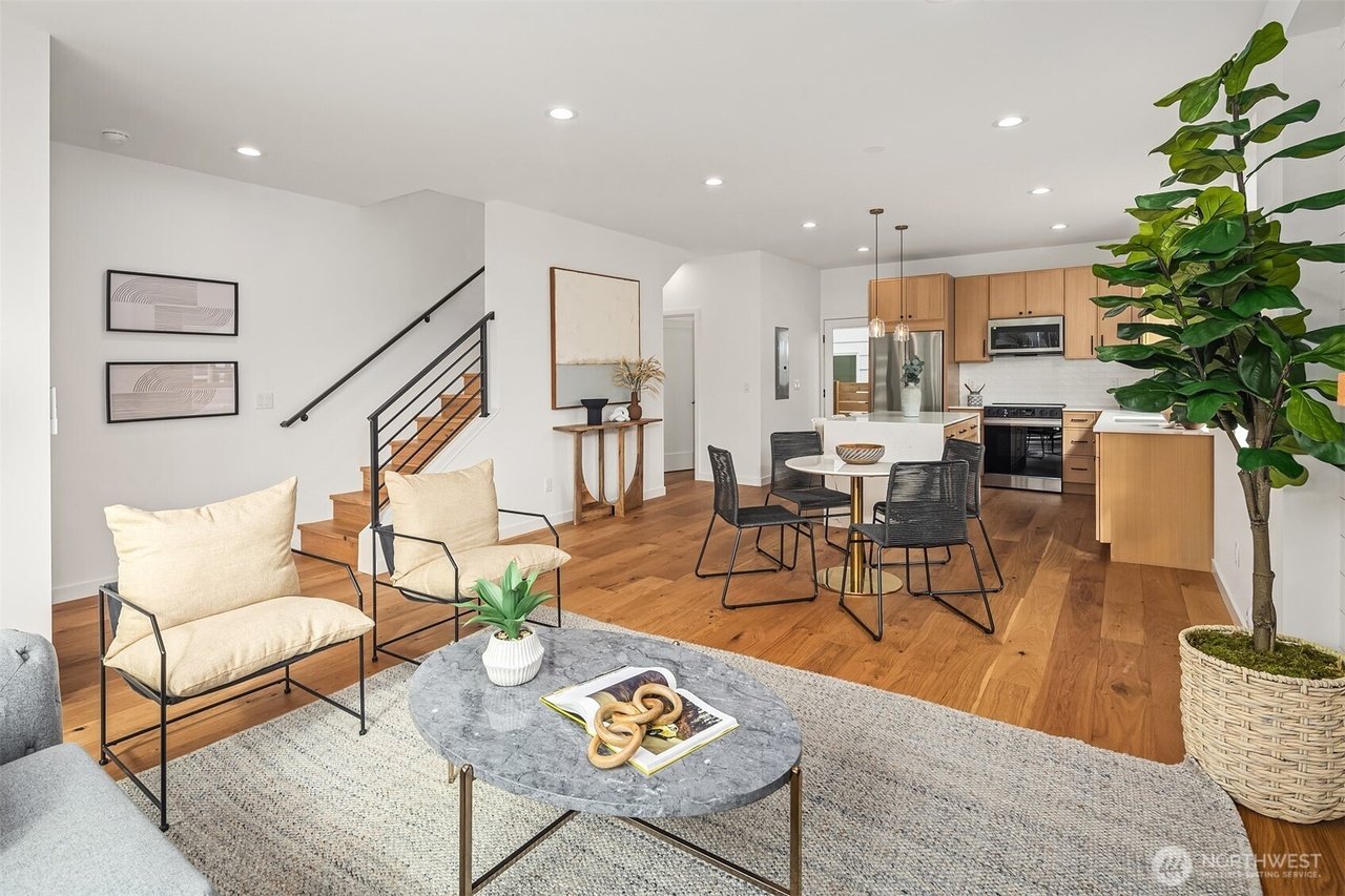 Modern open-plan living space with wooden floors, featuring a kitchen, dining area, and seating. The kitchen includes wooden cabinets and stainless steel appliances. The dining area has a round table with black chairs. A large potted plant and minima