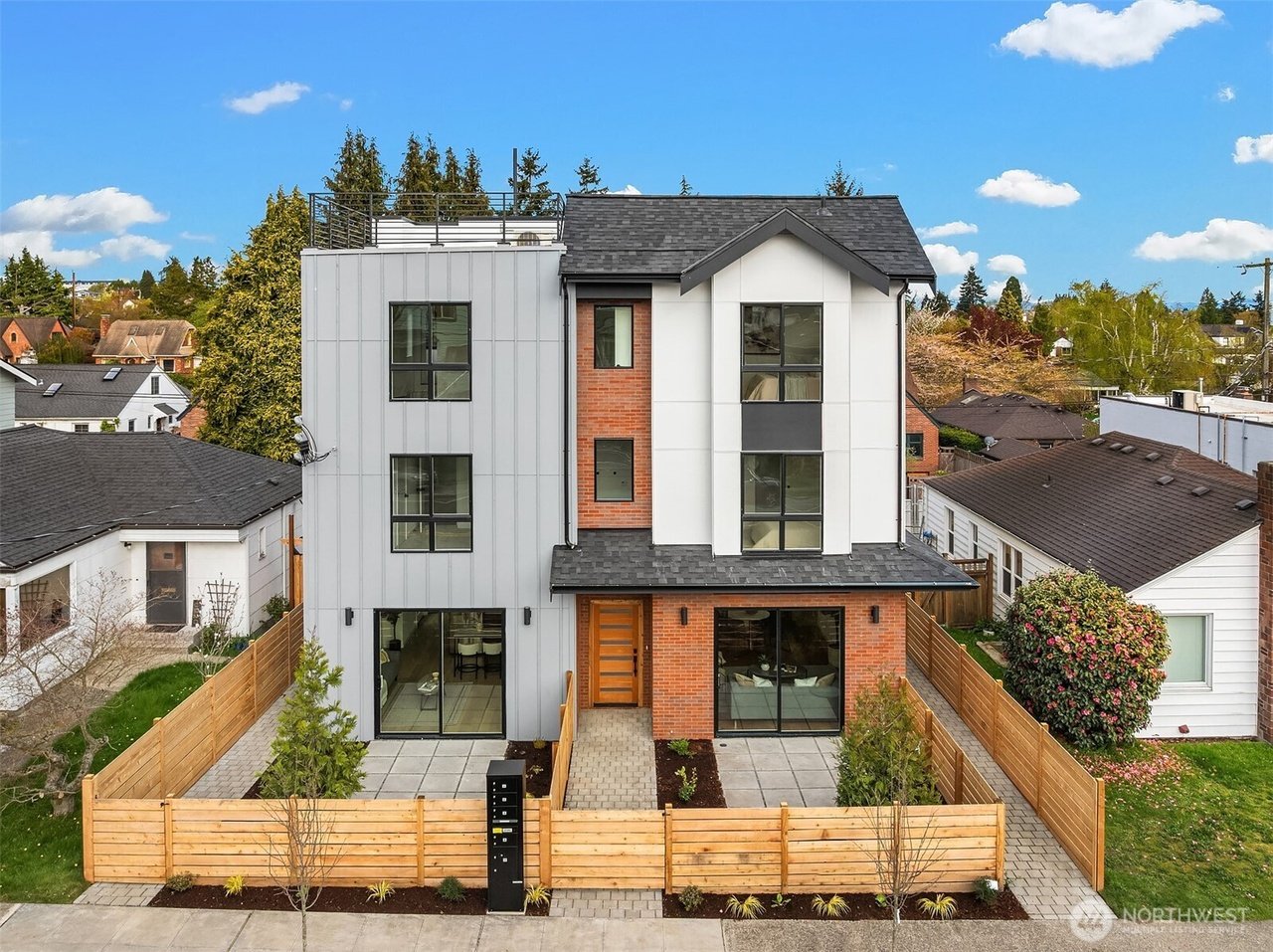Modern multi-story residential building with a mix of brick and metal siding, large windows, and landscaped front yard, surrounded by a wooden fence. Other houses and trees are visible in the background.