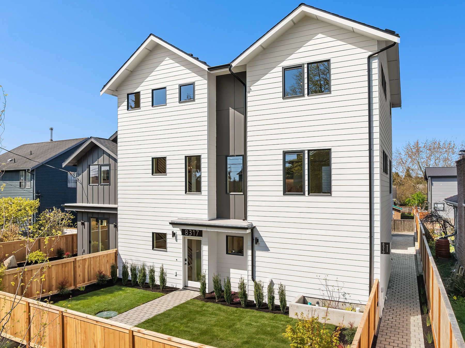White modern duplex house with two peaked roofs, multiple windows, and a wooden fence surrounding a landscaped front yard under clear blue sky.