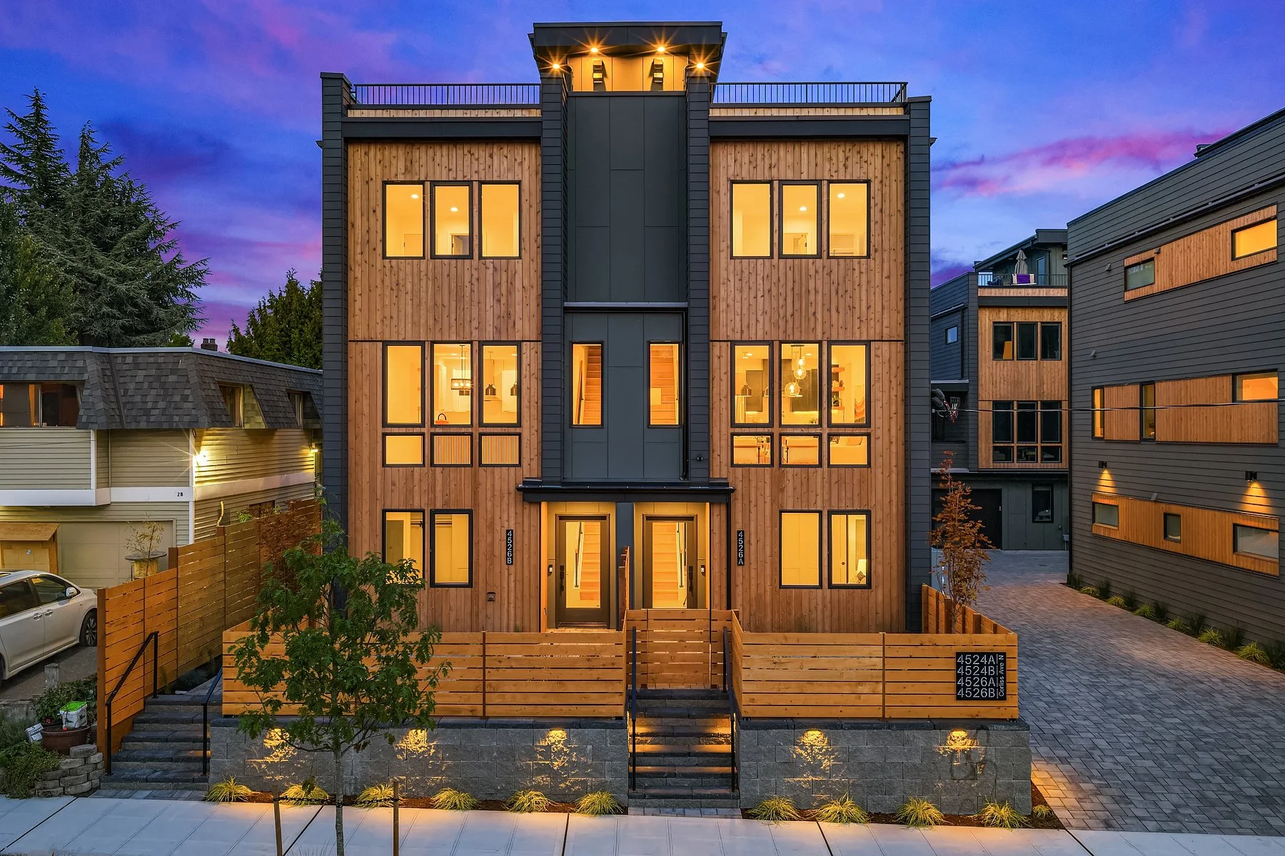 Modern multi-story house with large windows, wooden siding, and exterior lighting at dusk, surrounded by a wooden fence and neighboring houses.