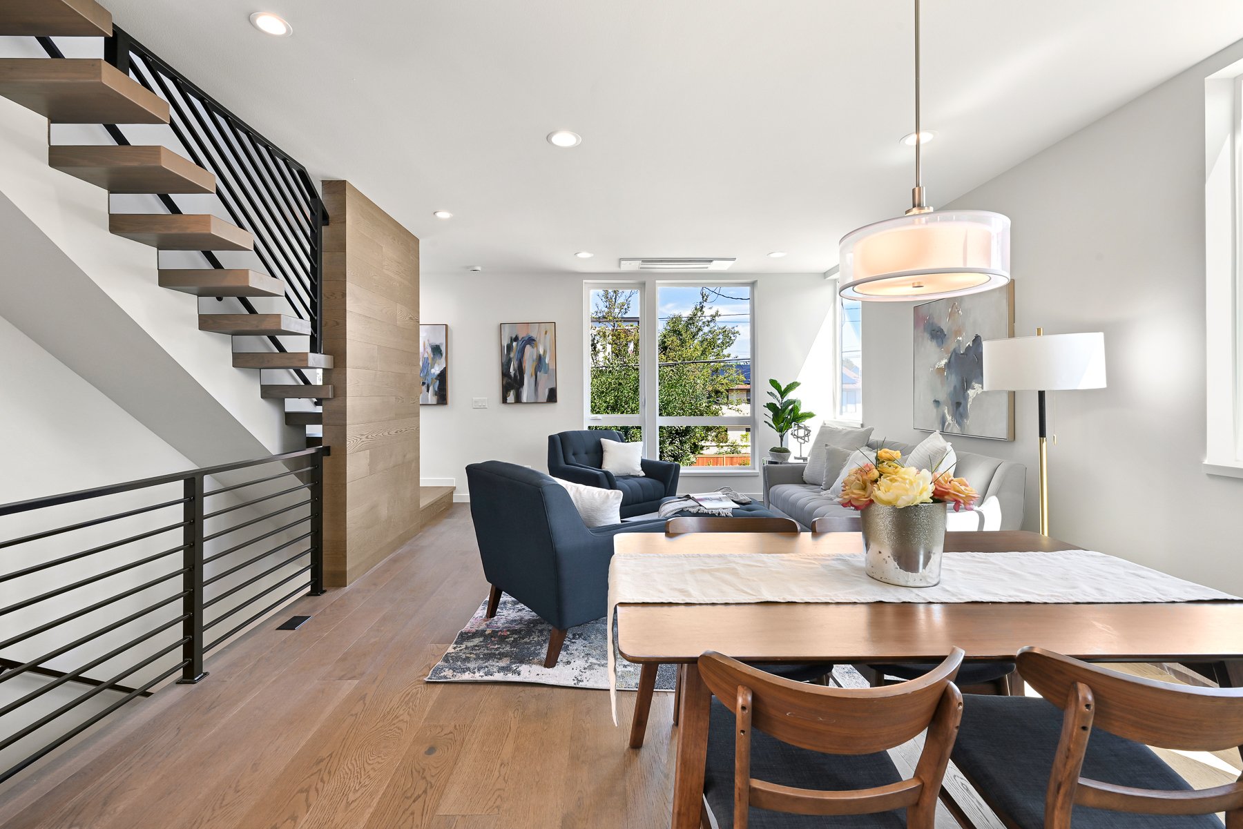 Modern living room and dining area with wooden staircase, light wood flooring, contemporary furniture, large windows, and wall art.