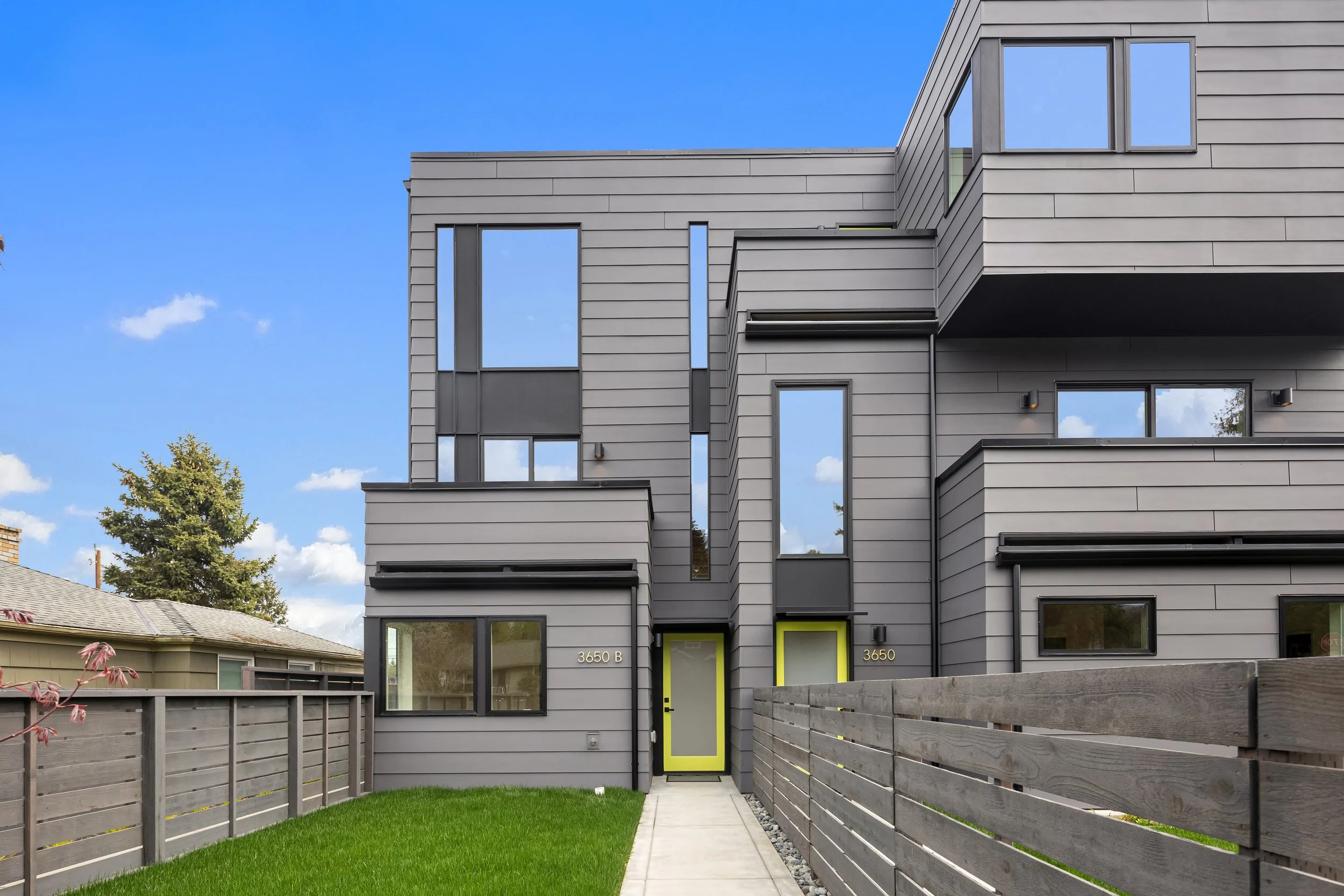 Modern gray multi-story apartment building with large windows, yellow door frames, and a front yard with a concrete walkway and wooden fence under a partly cloudy sky.