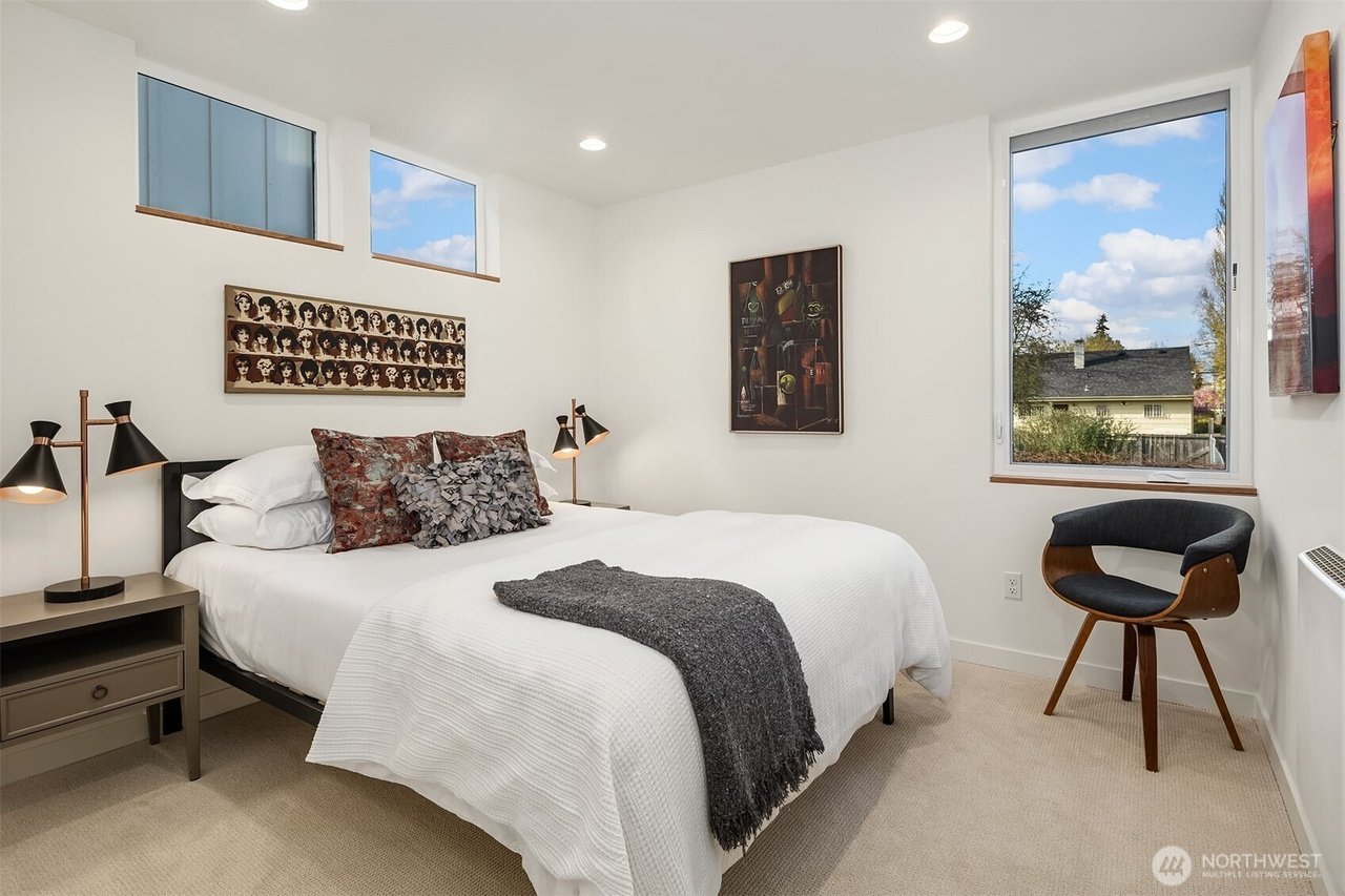 Modern bedroom with white bedding, decorative pillows, and a blanket, two black table lamps on nightstands, a dark wood chair, and artwork on the walls. Large windows provide natural light.