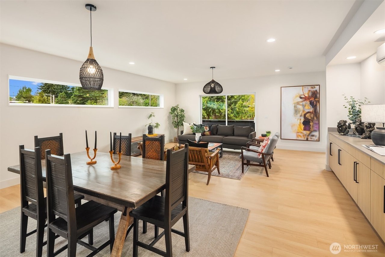 Modern living and dining area with wooden table, black chairs, contemporary lighting, sofa, armchairs, large windows, abstract wall art, and decorative plants.