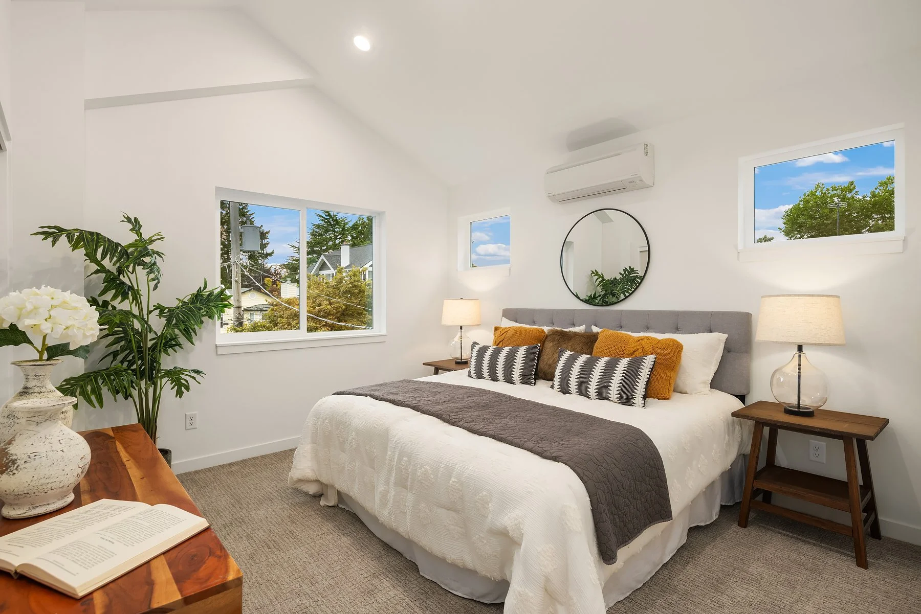 A cozy bedroom with white walls, a large bed with gray, brown, and white pillows, and two wooden nightstands with lamps. There are three windows showing trees and a blue sky, a round mirror on the wall, air conditioning unit, a potted plant, a wooden