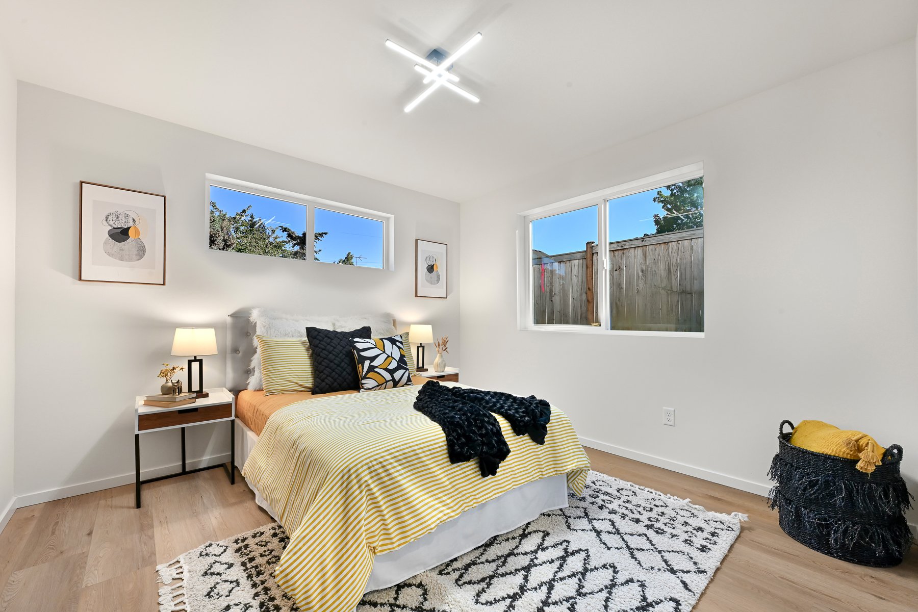 Modern bedroom with white walls, wooden floor, bed with yellow and white striped bedding, two nightstands with lamps, abstract artwork on walls, geometric-patterned rug, and basket with folded blankets.