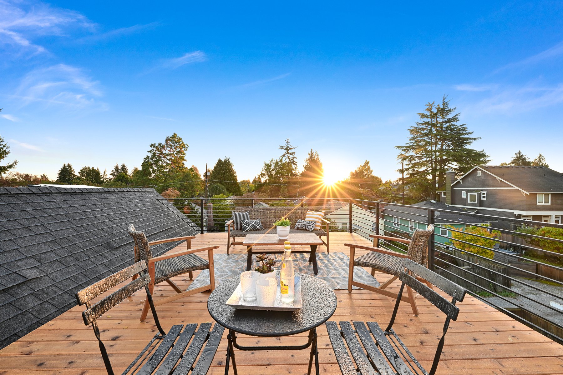 Rooftop deck with outdoor seating and a view of trees and rooftops, featuring a small table with drinks and a tray, under a clear blue sky with a setting sun.