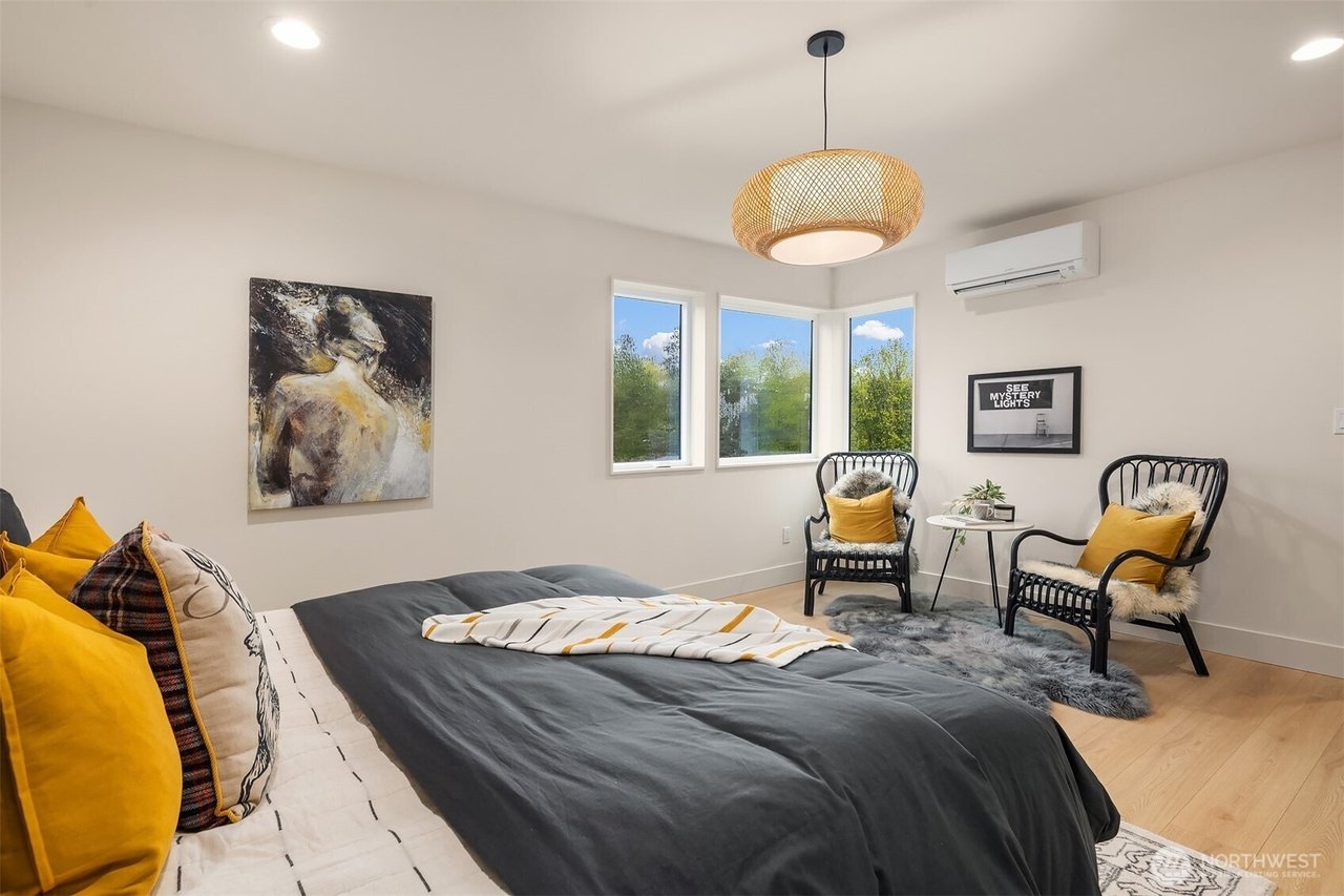 Modern bedroom with a bed featuring gray and yellow pillows, a painting on the wall, a pendant light, a small sitting area with two black chairs and a round table, light wood flooring, and large windows with greenery outside.
