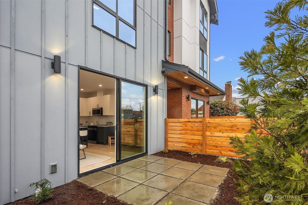 Modern residential building exterior with a small patio. Features a sliding glass door leading to an interior kitchen area, a wooden privacy fence, and outdoor lighting. Surrounded by neatly landscaped plants and trees.