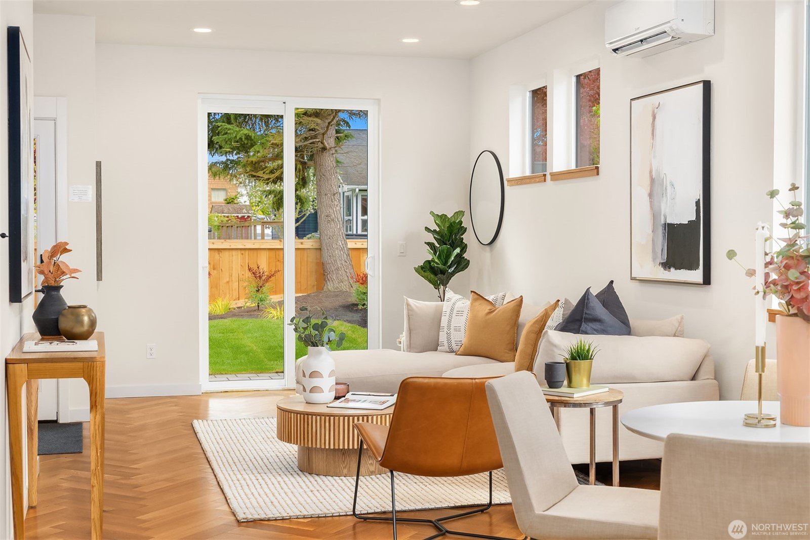 Modern living room with light beige walls, a white sofa with assorted pillows, brown leather armchair, round wooden coffee table, and sliding glass doors leading to a backyard with a wooden fence and tree. Artwork and indoor plants decorate the space