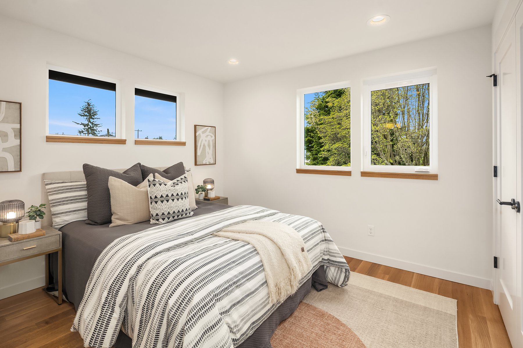 Modern bedroom with a neatly made bed, featuring striped bedding and decorative pillows. Three windows on the wall with views of trees, two wooden nightstands with lamps and plants, and abstract art on the walls. Light wooden floor and white walls.