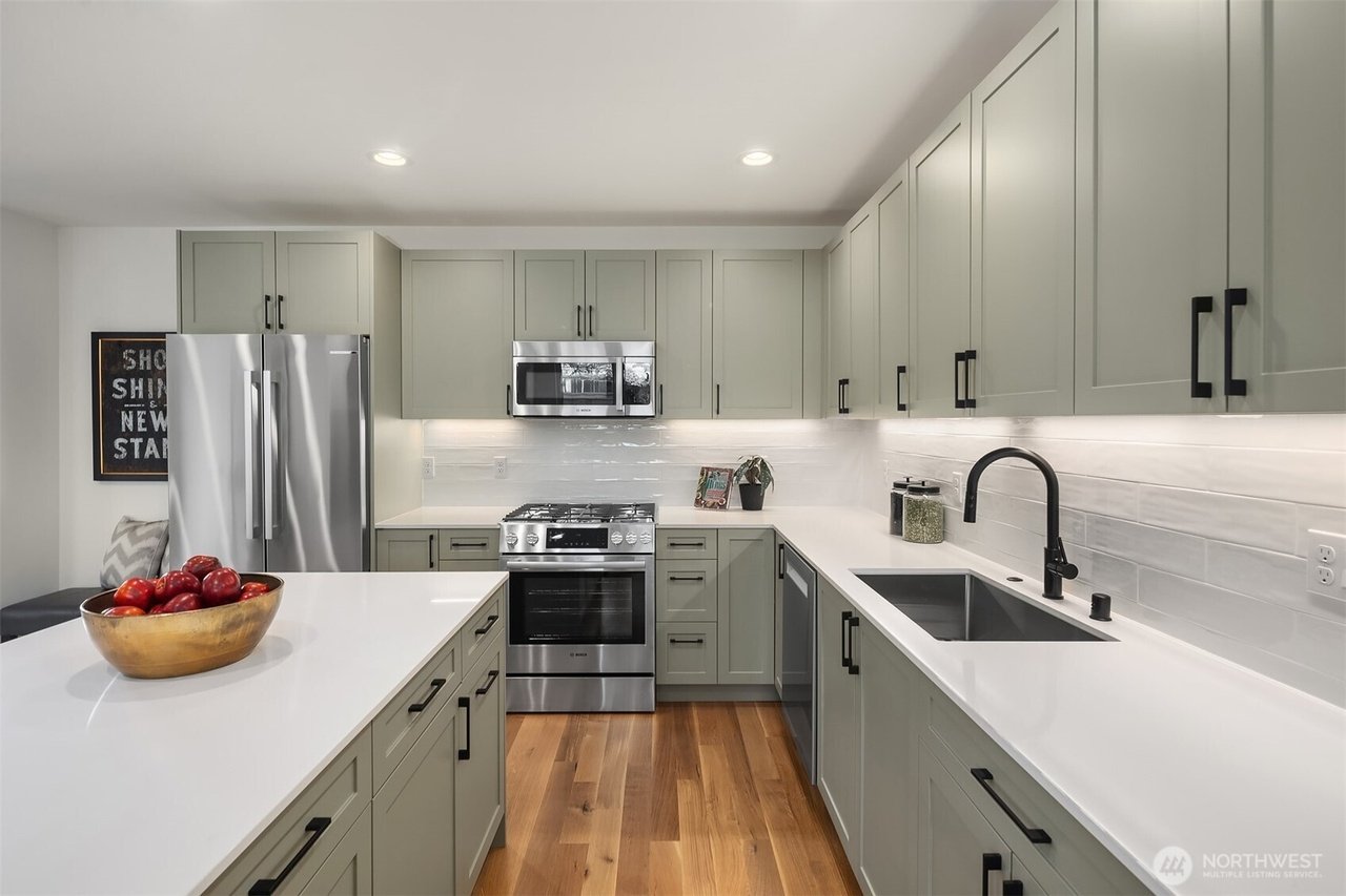 Modern kitchen with light green cabinets, stainless steel appliances, and a white countertop. A bowl of red apples sits on the island, and a black faucet is over the sink. The kitchen features a wooden floor and subtle under-cabinet lighting.