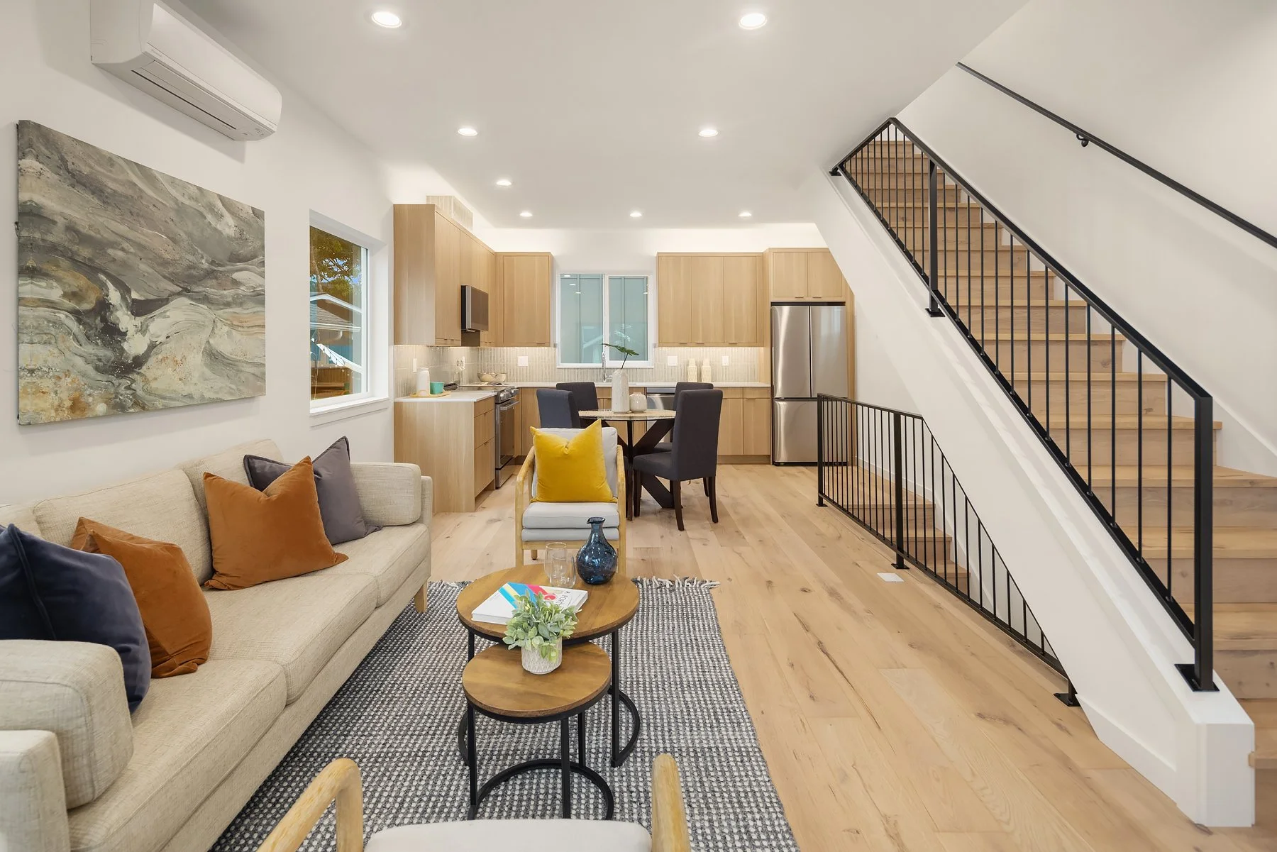 Open-concept living room and kitchen with wooden flooring, beige sofa with throw pillows, a small coffee table, a dining table with chairs, and a staircase with black railing leading upstairs.