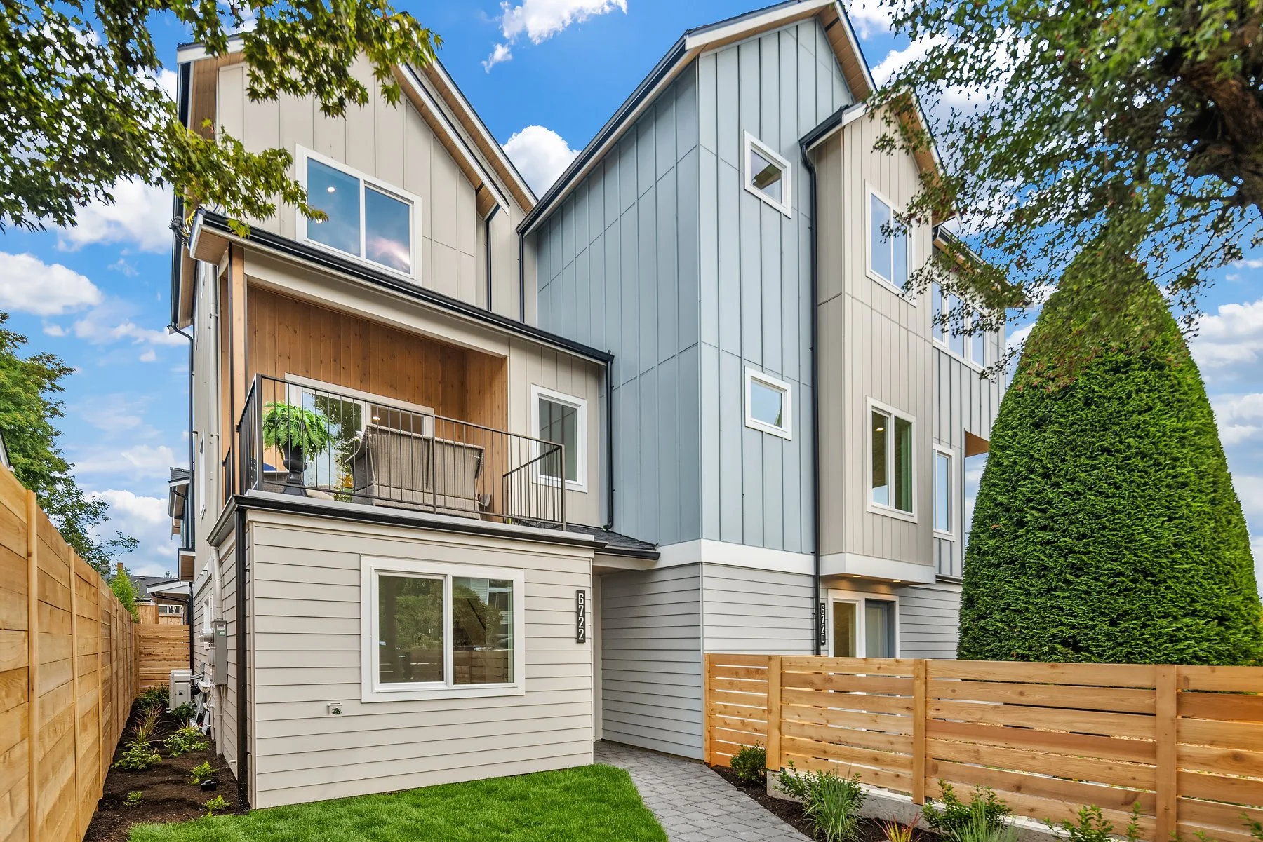 Modern multi-story residential building with beige and blue siding, balcony with potted plants, large windows, surrounded by a wooden fence, green lawn, trees, and a partly cloudy sky.