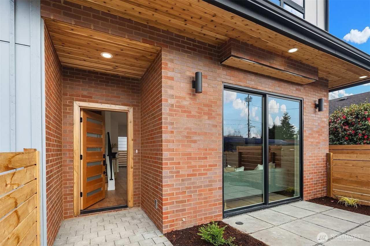 Modern brick house exterior with open wooden door, large glass window, and patio.