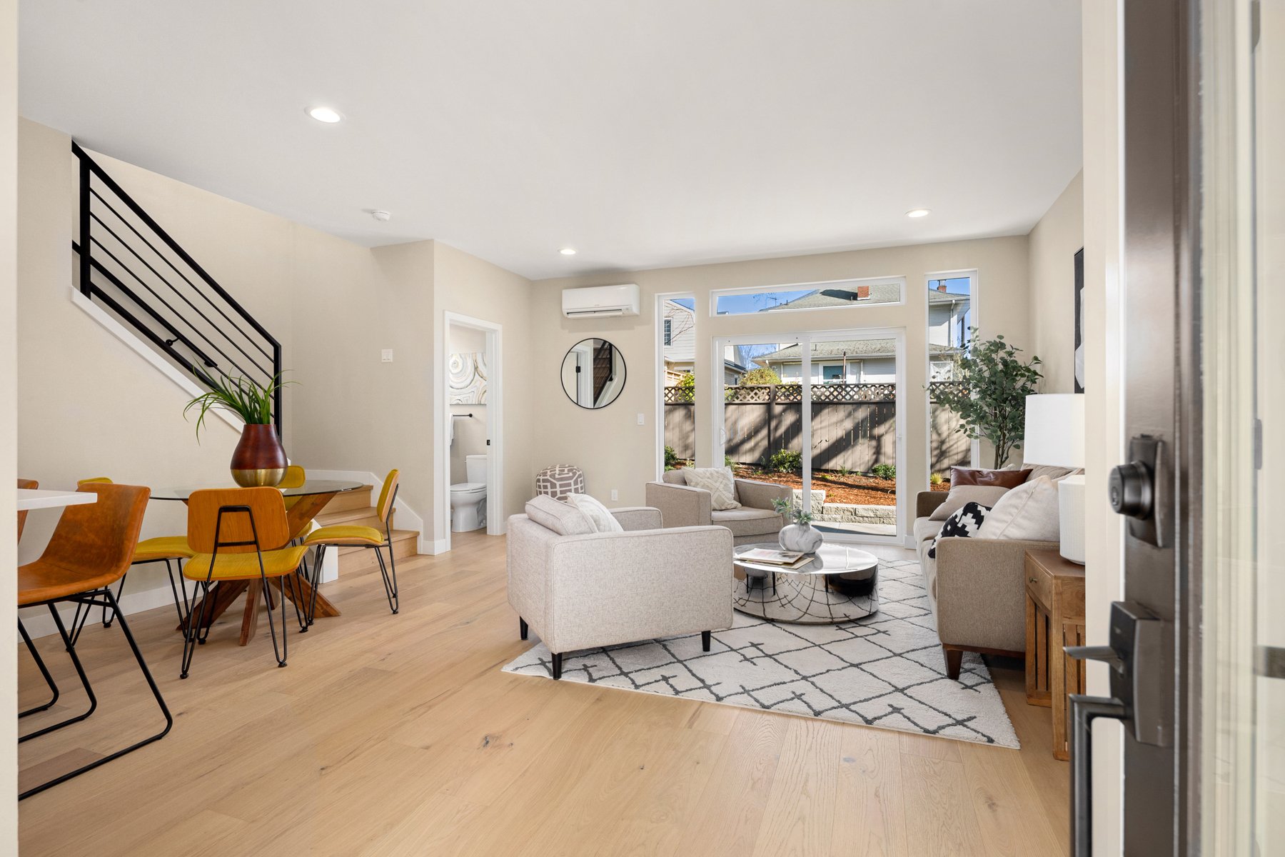 Modern living room and dining area with beige walls and light wood flooring, featuring a gray sofa set, glass coffee table, and a dining table with yellow chairs, with a view of a fenced backyard through large windows.