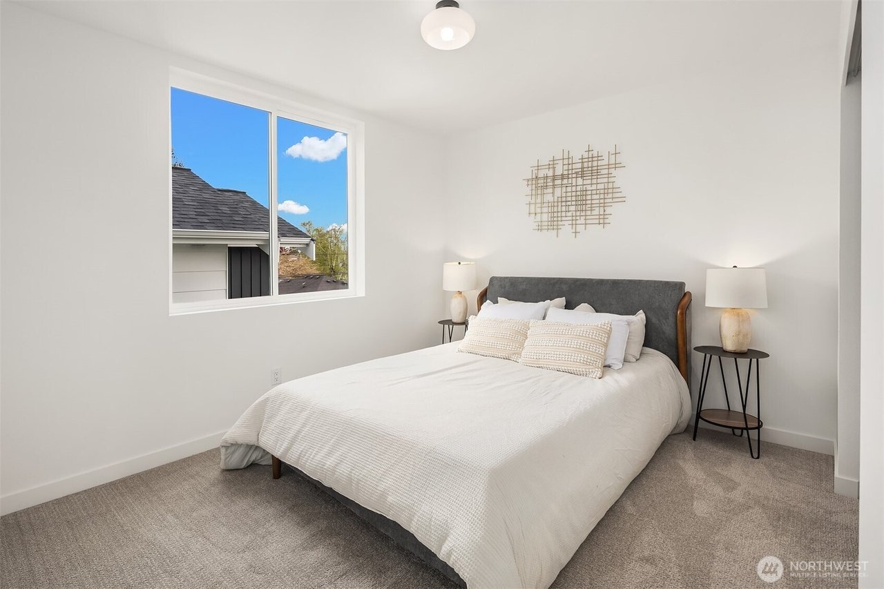Minimalist bedroom with a neatly made bed, decorative wall art, bedside tables with lamps, and a window with a view of a roof and sky.