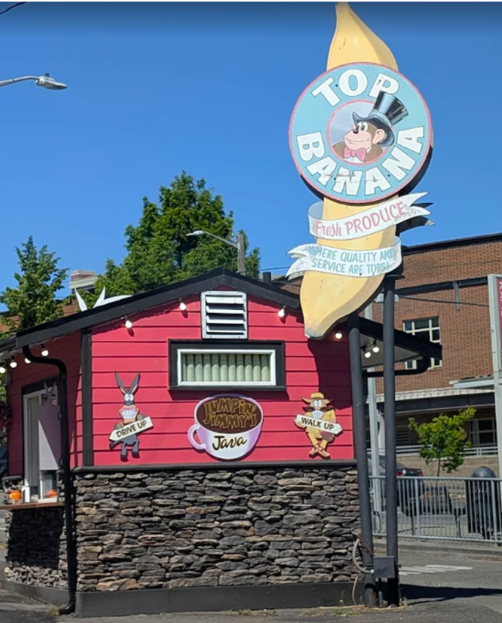Colorful sign for Top Banana Fresh Produce with cartoon characters, including a rabbit and a man with a hat, outside a pink building with stone foundation, under a clear blue sky.