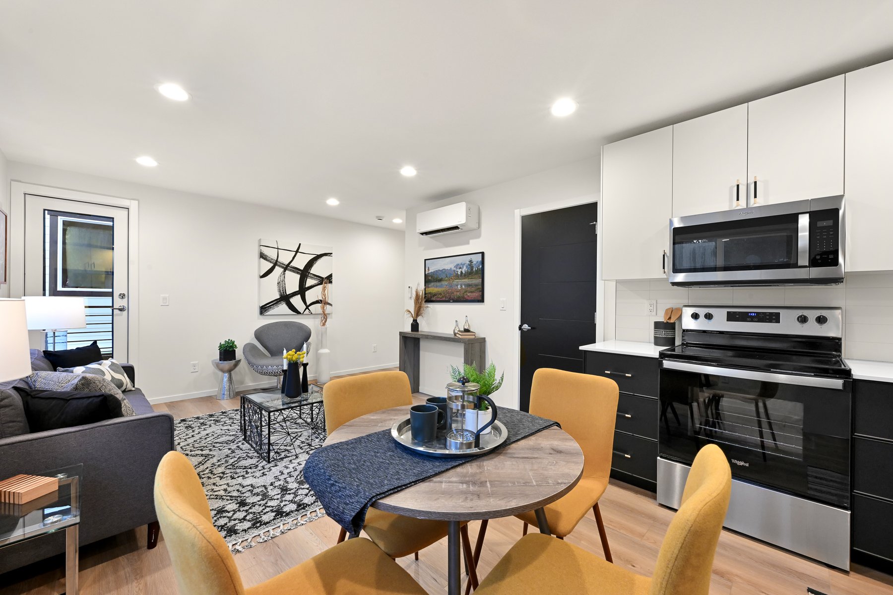 Open-concept living and kitchen area with modern decor, a dining table set with yellow chairs, a gray couch, abstract art, and stainless steel appliances.