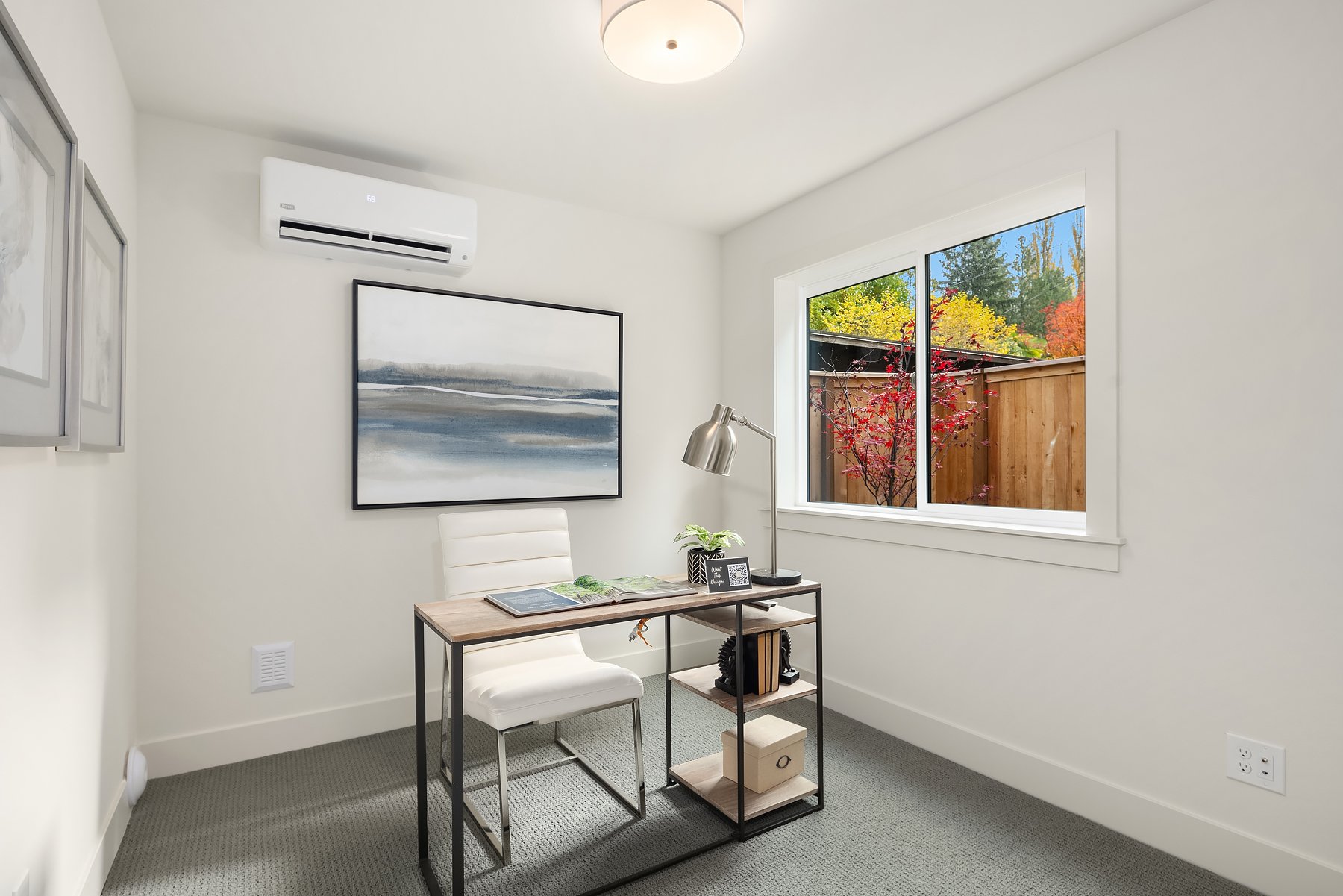 Minimalist home office with a desk, white chair, and a desk lamp. A large framed abstract artwork hangs on the wall, and a window displays a view of colorful autumn foliage. Room includes an air conditioning unit and neutral decor.