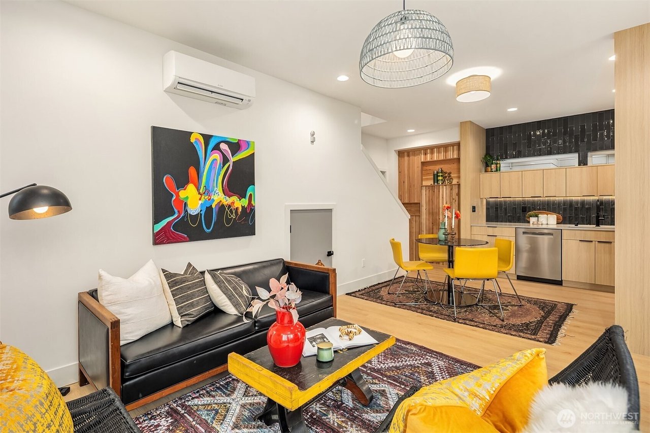 Modern living room and kitchen with black leather couch, colorful artwork, yellow chairs, light wood cabinetry, and stainless steel appliances.
