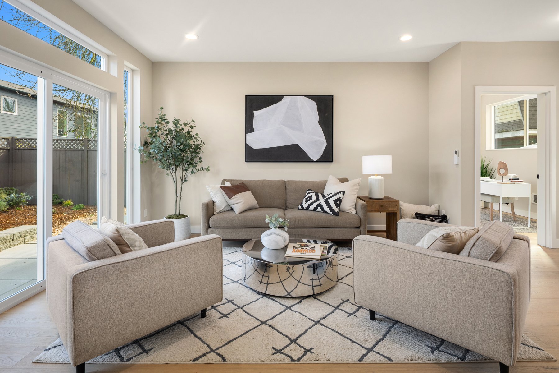 Modern living room with beige sofas, abstract wall art, and a round coffee table on a patterned rug. Large windows provide natural light, and a potted plant adds greenery. A side table with a lamp is beside the sofa."