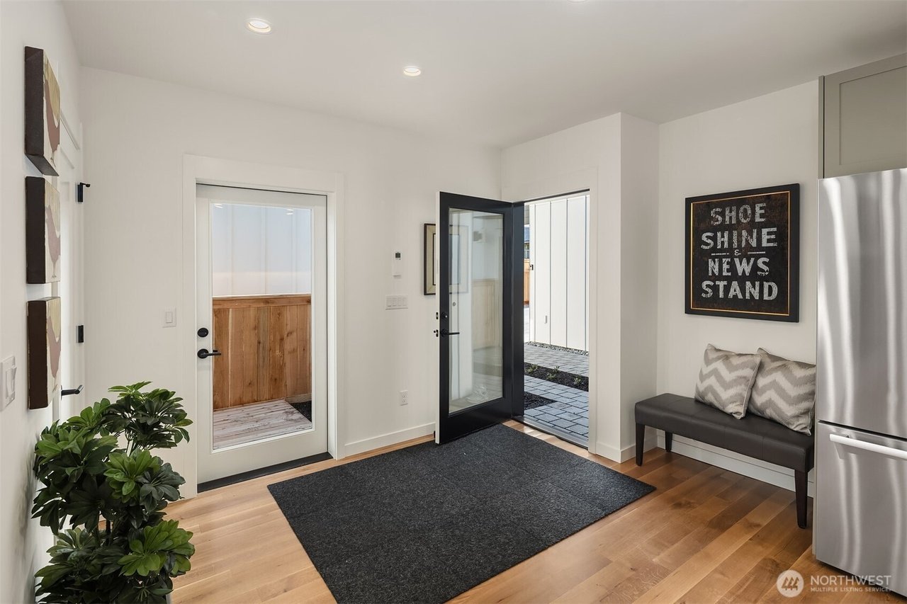 Modern entryway with glass door, indoor plant, bench with cushions, hardwood floor, and wall art reading "Shoe Shine & News Stand."