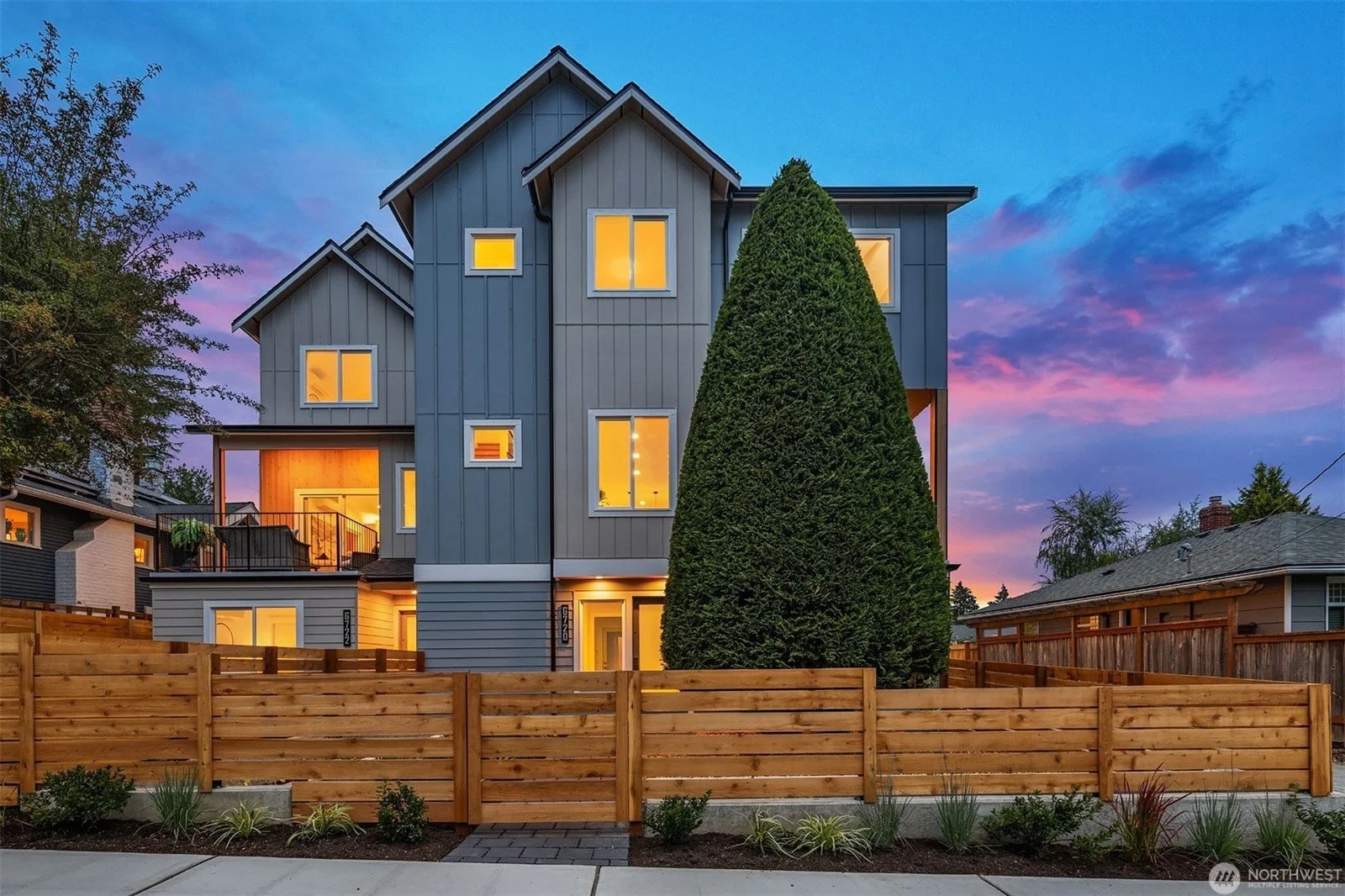 Modern multi-story house with gray exterior walls, large and illuminated windows, a sharp roofline, and a tall privacy hedge in front, fenced with a wooden fence, at dusk with a colorful sky.