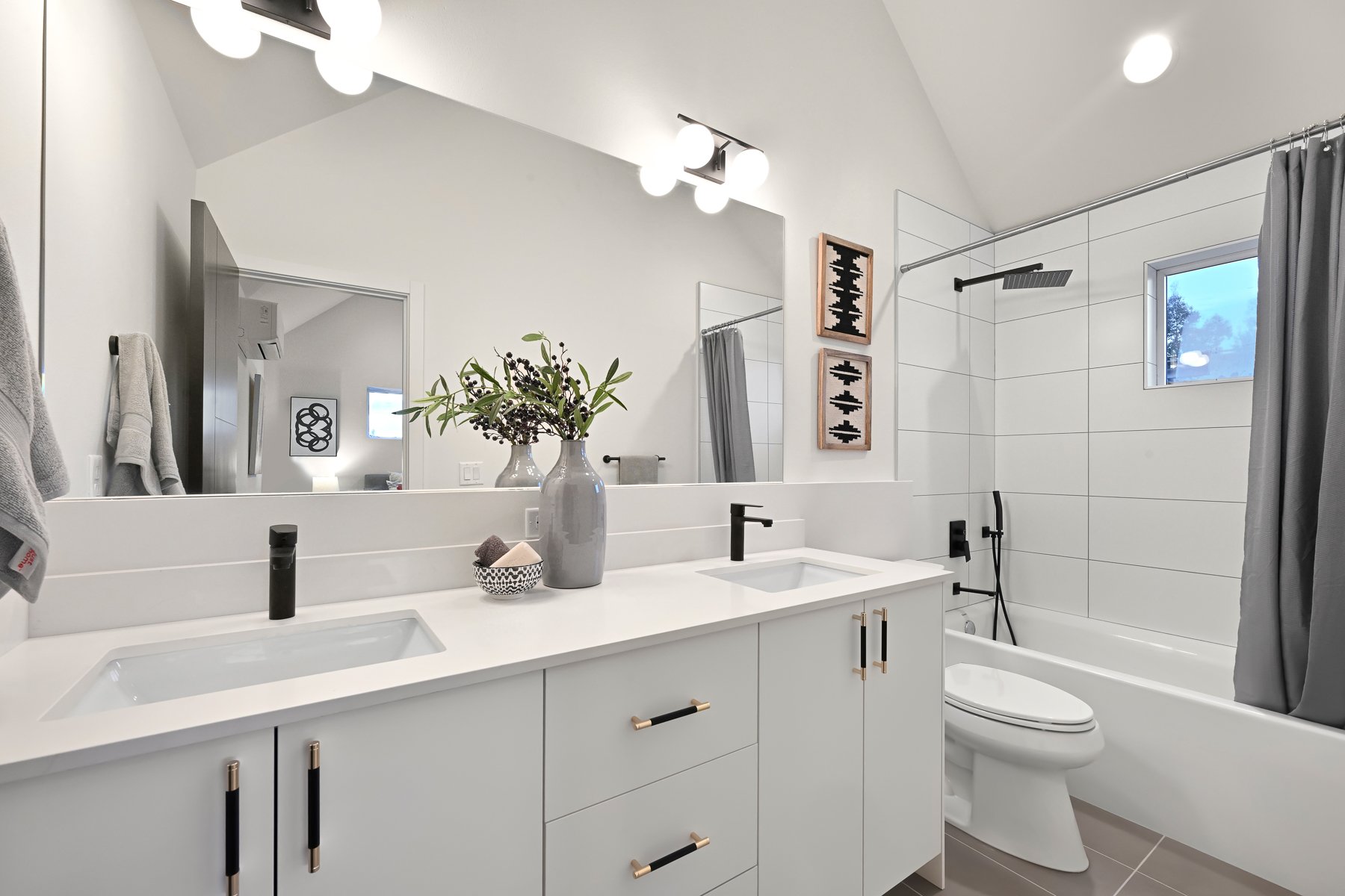 Modern bathroom with white cabinets, double sinks, large mirror, decorative vase with greenery, wall art, toilet, bathtub, and shower curtain.