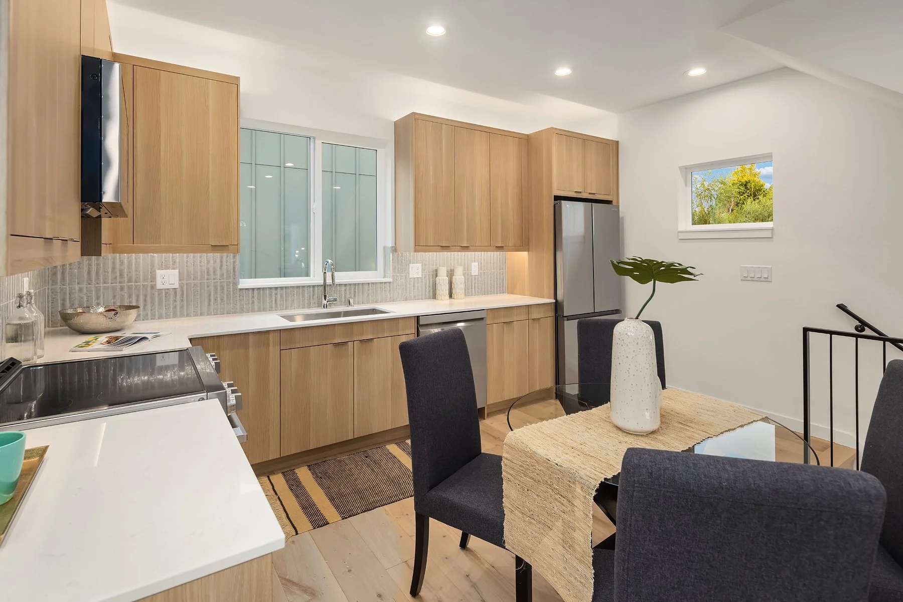 Modern kitchen with wood cabinets, stainless steel refrigerator, and a dining table with a beige runner and dark chairs, decorated with a white vase and a large green leaf.