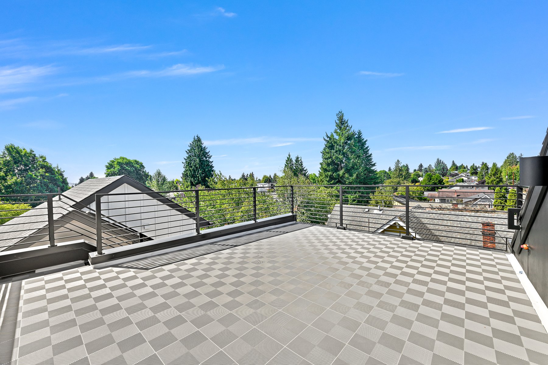 Rooftop terrace with checkered flooring and metal railing, overlooking a suburban neighborhood with trees and rooftops under a clear blue sky.