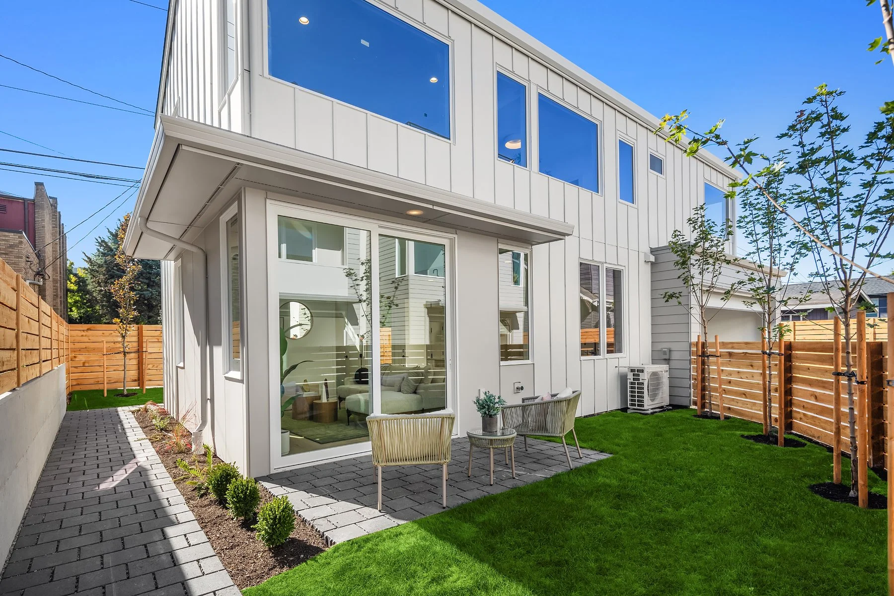 Modern two-story house with white exterior, large windows, patio with chairs and potted plant, green lawn, young trees, wooden privacy fence, clear blue sky