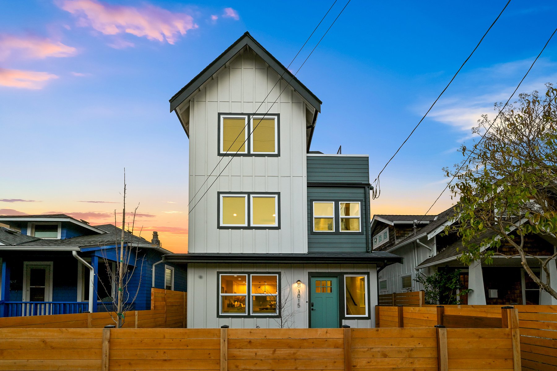 Modern three-story house with white and gray exterior, a teal front door, and wooden fence at sunrise.
