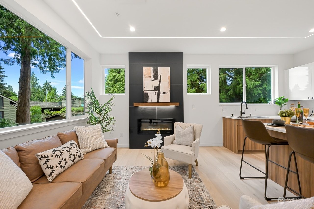 Modern living room with brown leather sofa, patterned throw pillows, a round coffee table with a vase, and a chair. A fireplace, abstract artwork, and large windows with a view of trees are visible. Elegant open kitchen with bar stools can be seen.