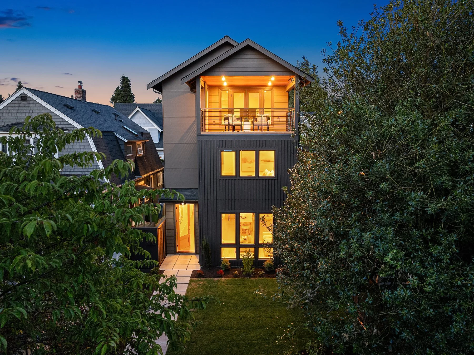 A modern multi-story house illuminated at dusk with outdoor lighting, featuring large windows, a balcony on the upper floor, and surrounded by greenery and neighboring houses.