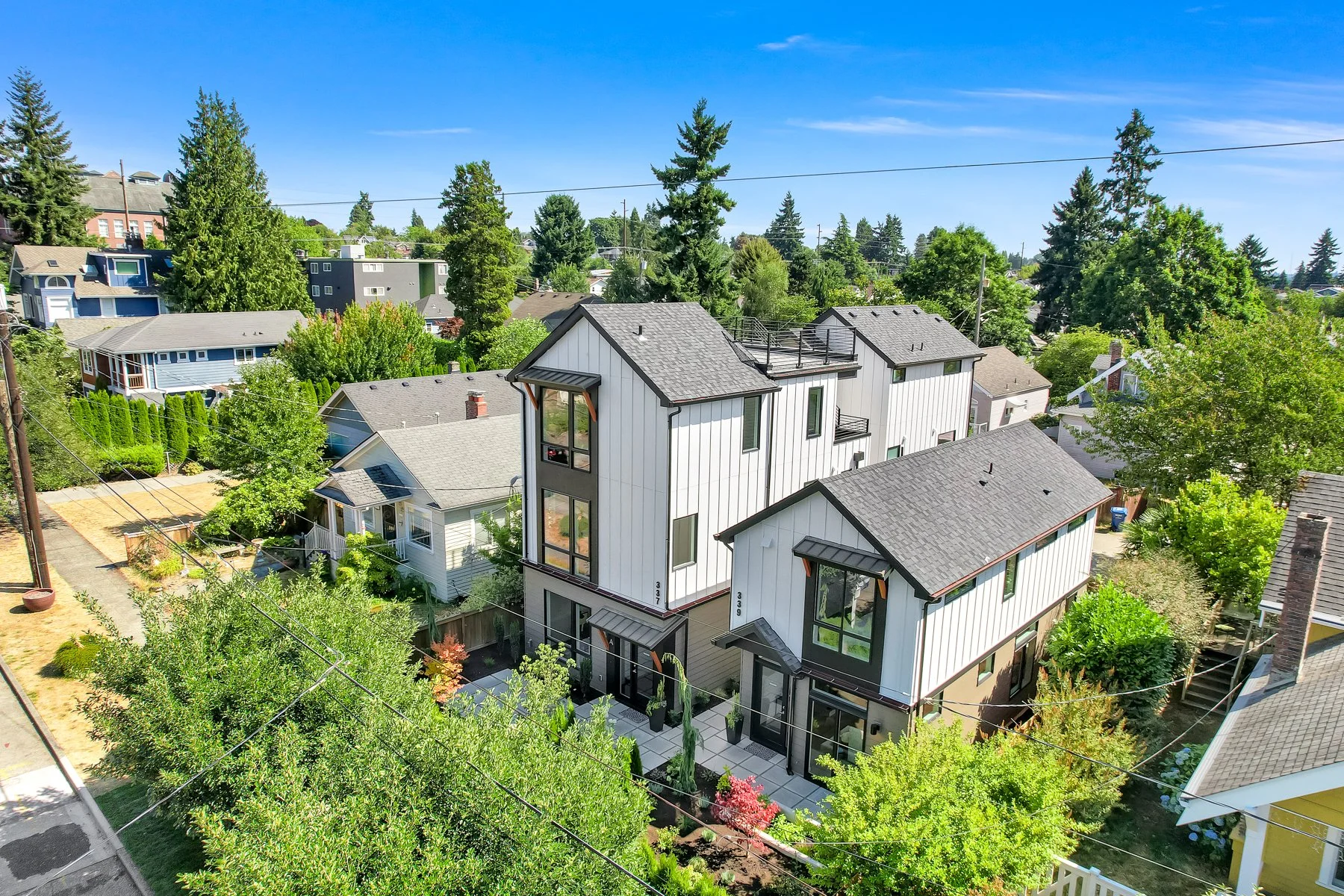 Aerial view of modern residential houses with gray roofs, surrounded by greenery and trees, in a suburban neighborhood under a clear blue sky.