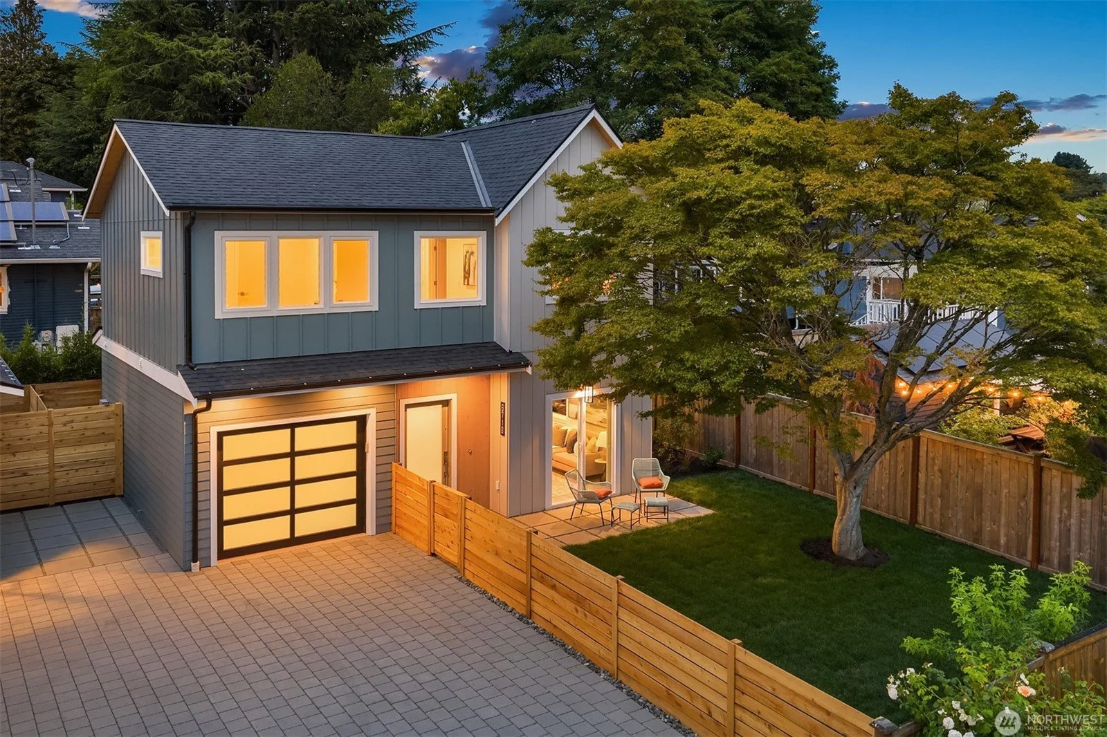 A modern, two-story house with a well-lit interior, a front porch with chairs, a driveway with pavers, a large tree, and a wooden fence in the backyard during evening.