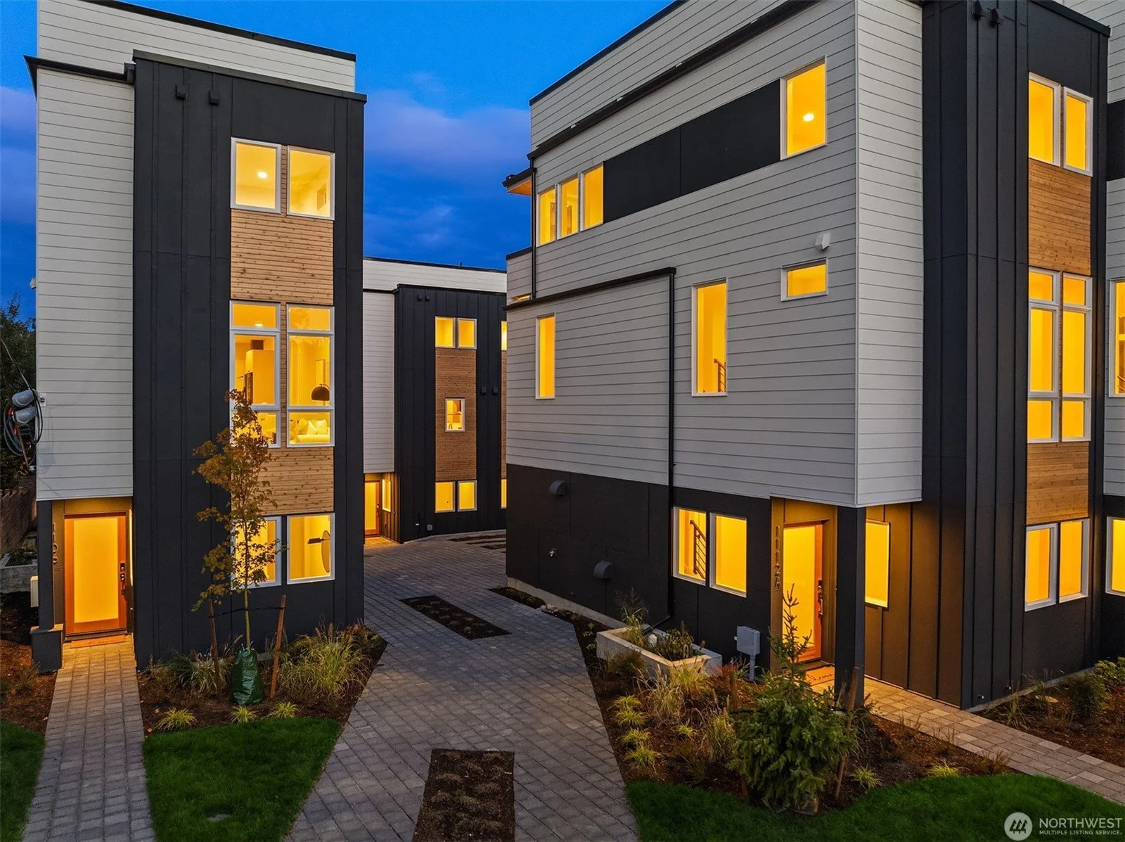 Modern multi-story apartment buildings with white, black, and wood siding, lit windows glowing in the evening, landscaped pathways, and trees in front.
