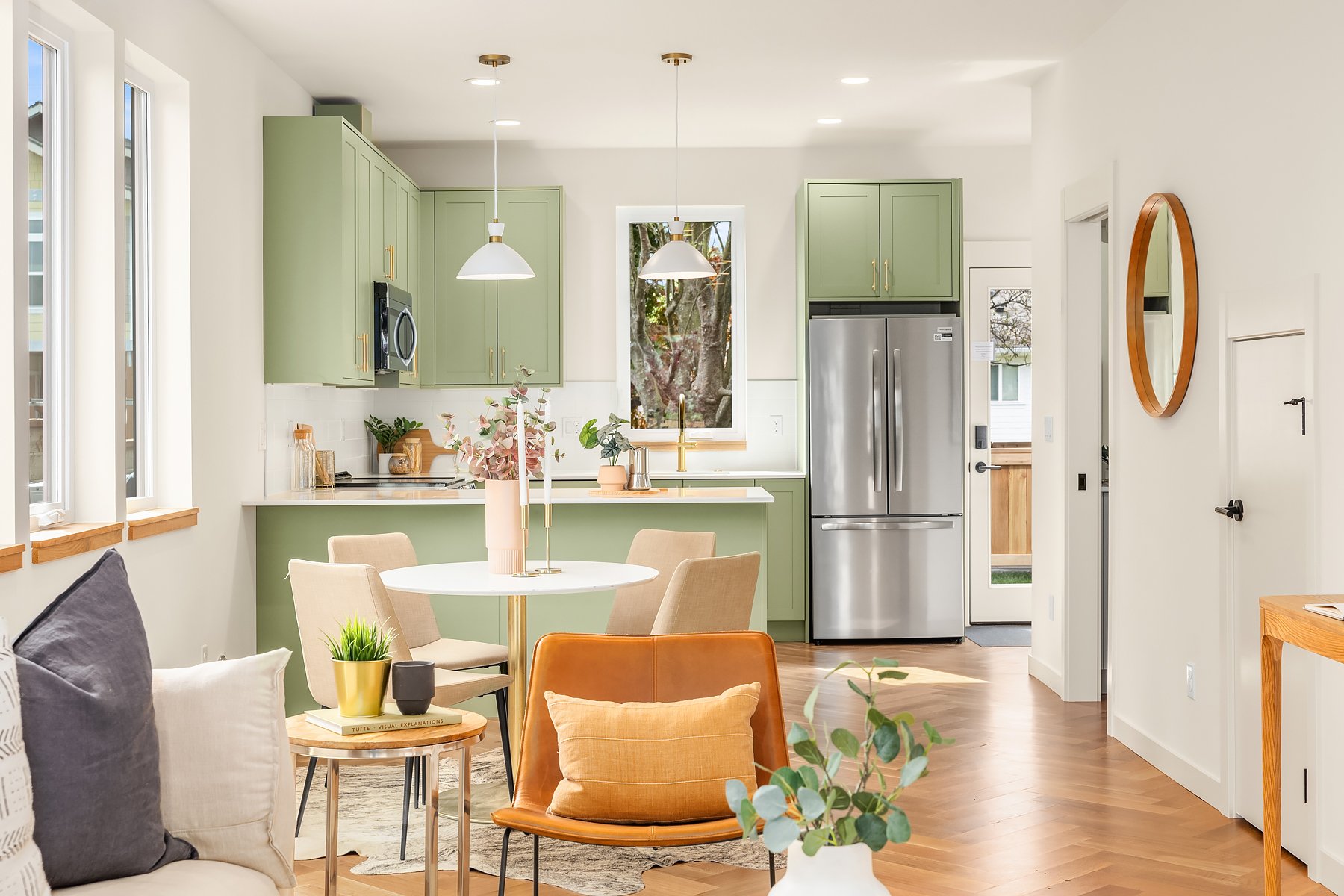 Modern kitchen and dining area with green cabinets, stainless steel appliances, a round white dining table, beige chairs, orange accent chair, and decorative plants. Light wood flooring and pendant lighting add warmth to the space.