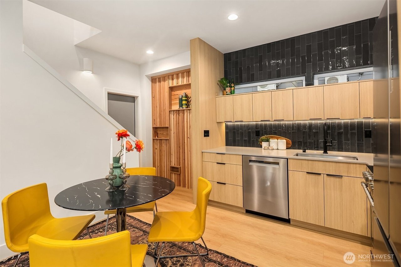 Modern kitchen and dining area with wood cabinetry, black backsplash, stainless steel appliances, a black marble round table with yellow chairs, and decorative flowers on the table.