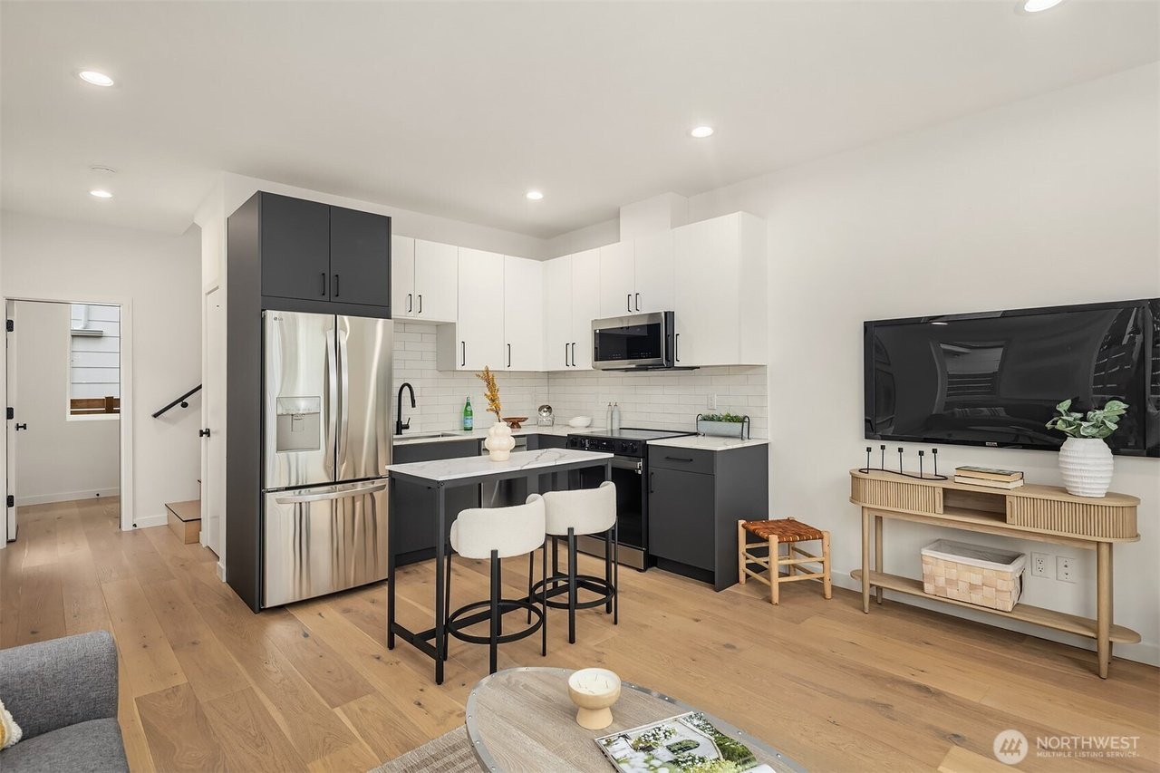 Modern kitchen and living area featuring a sleek design with stainless steel appliances, white and dark cabinets, a small island with two chairs, a wall-mounted TV, wooden flooring, and minimalistic decor.