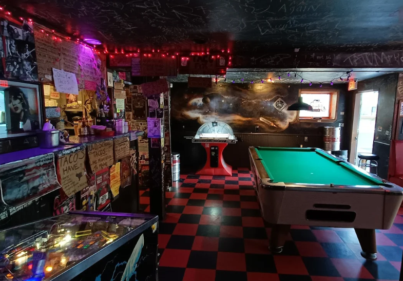 Interior of a gaming bar or arcade with a pool table, mini foosball table, ping pong table, arcade machine, and wall decorations.