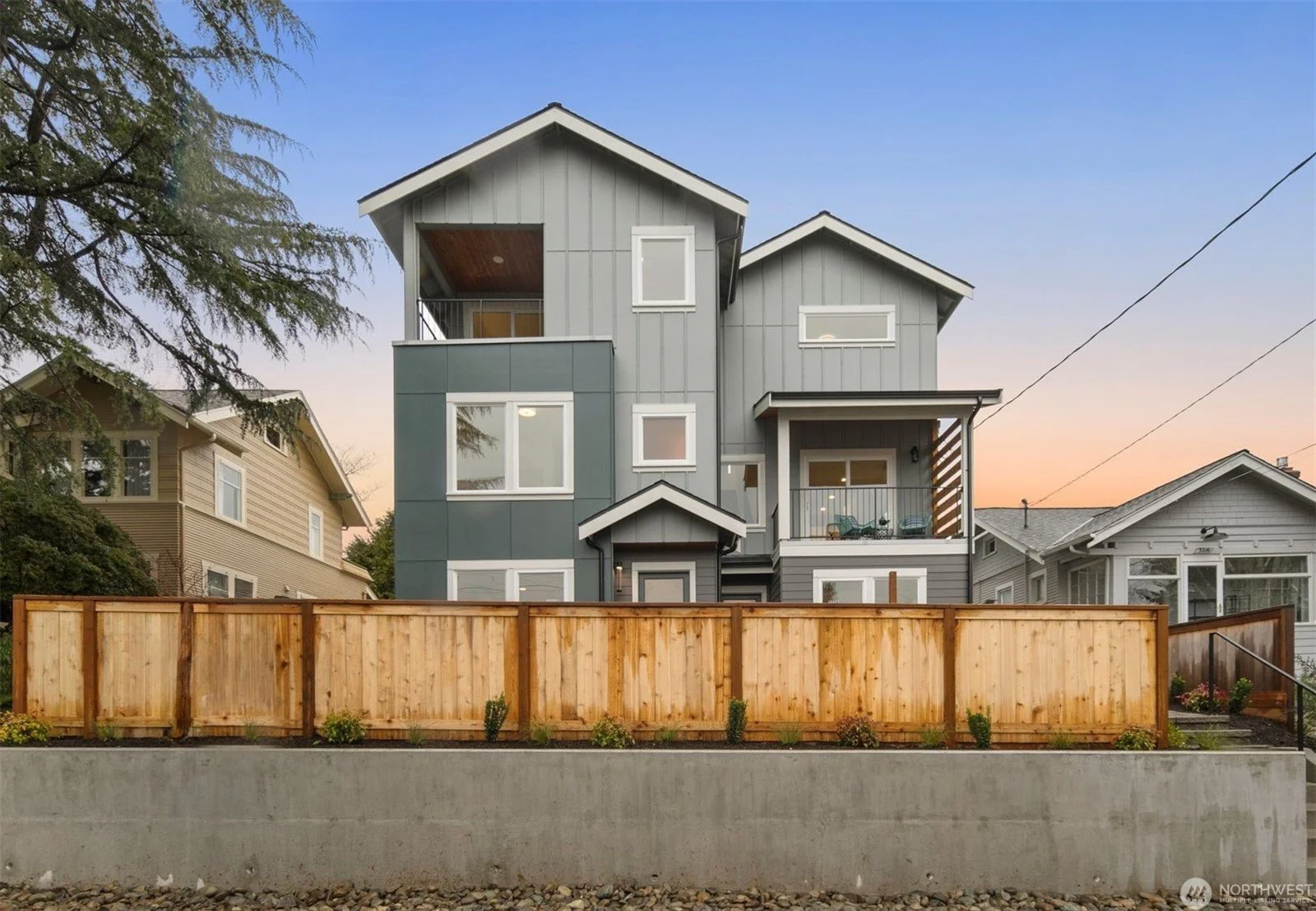 A modern, multi-story house with gray siding and large windows, surrounded by a wooden fence, with a neighboring house on each side, set against a sunset sky.