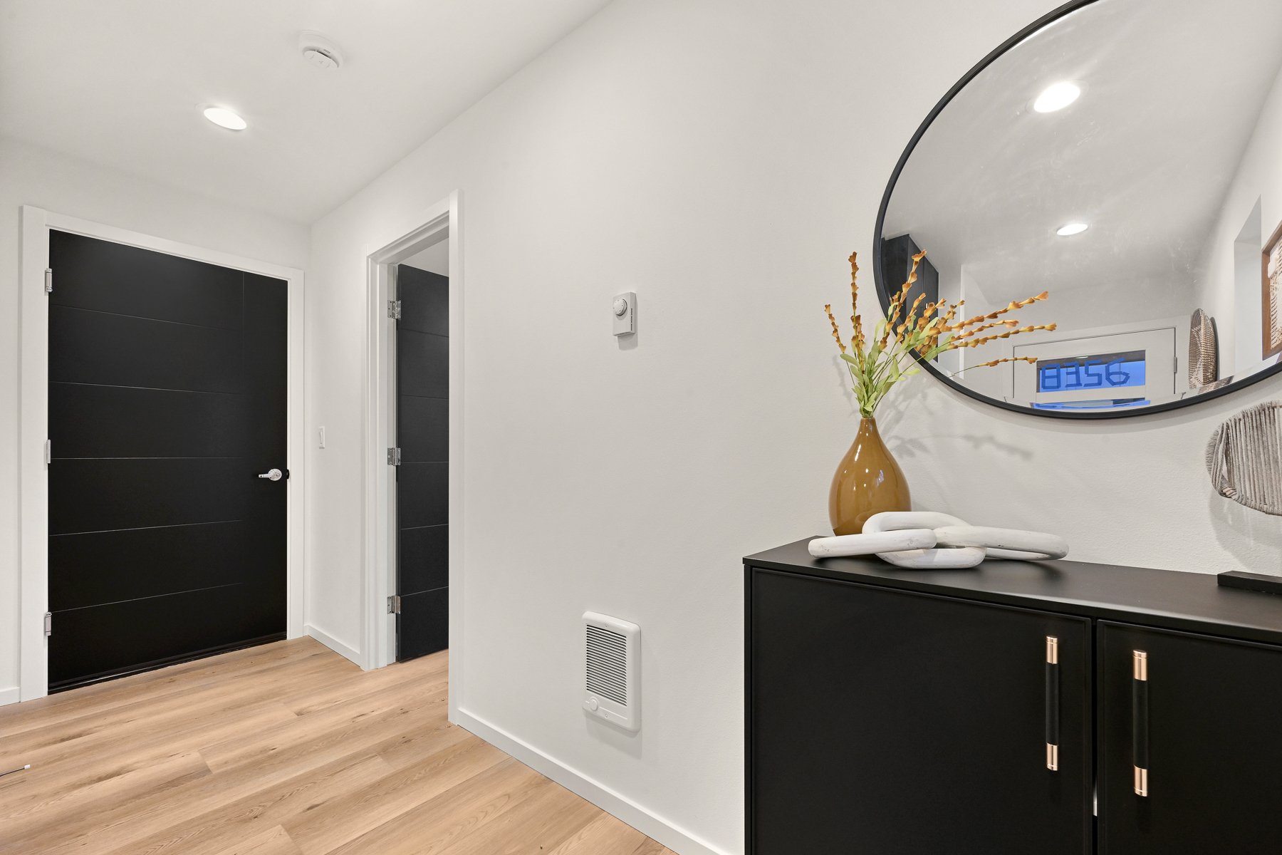 Modern hallway with black doors, light wood flooring, a round mirror, and a black cabinet with decorative items.