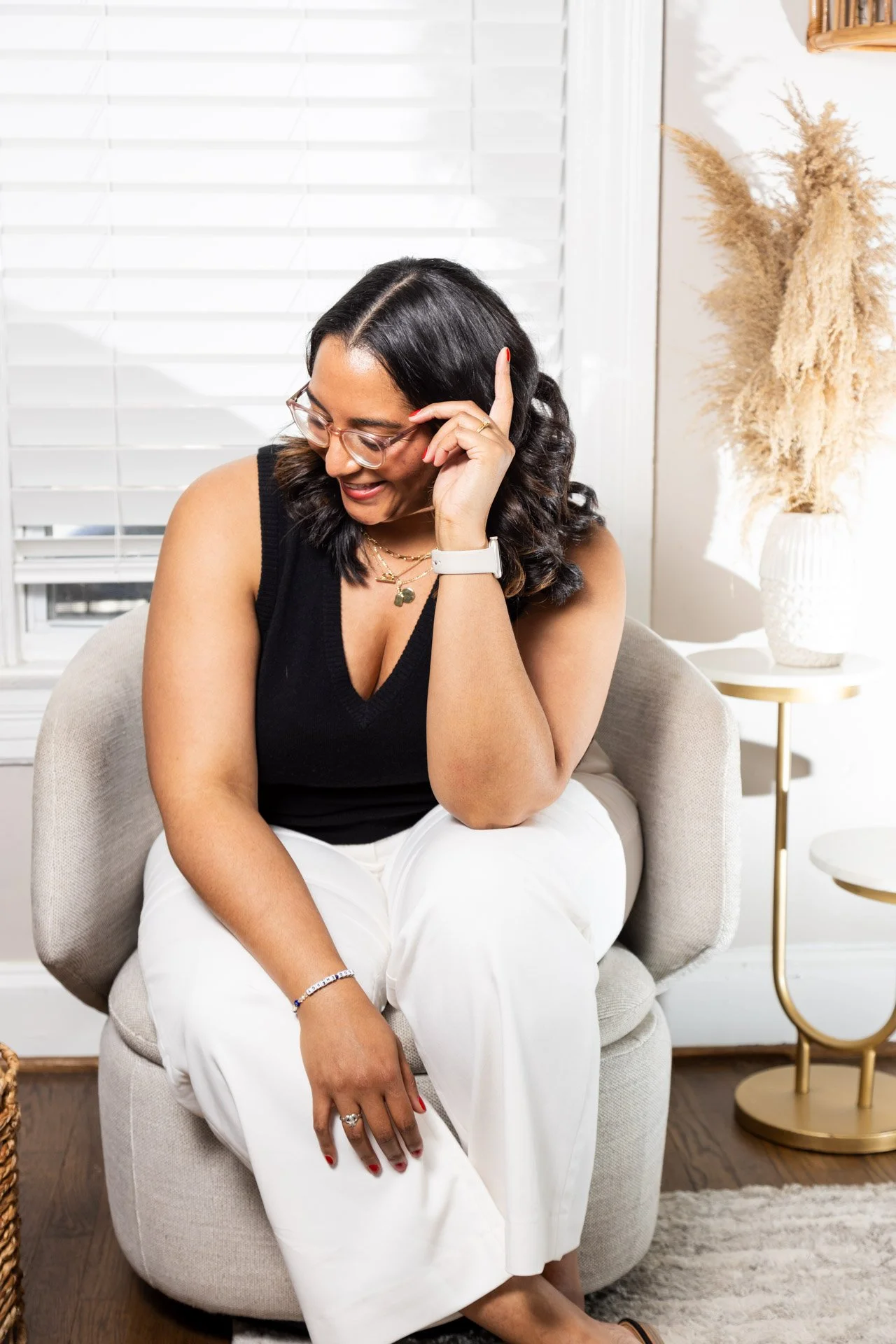 Renee sitting on a chair, wearing glasses, black top, and white pants, smiling and touching her head with her left hand, with home decor in the background.
