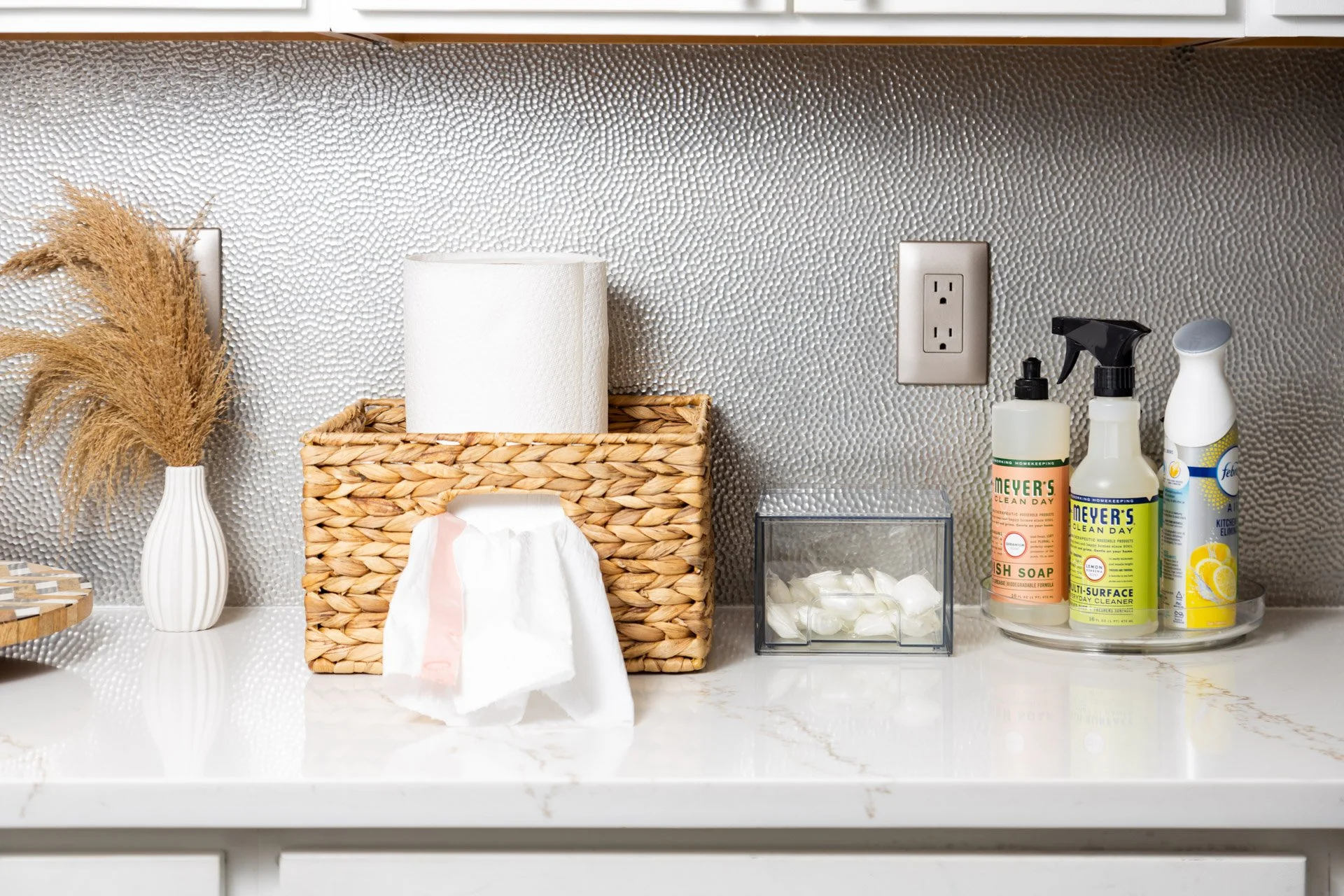 Countertop with cleaning supplies, paper towel holder, dried plant in vase, and textured silver backsplash.