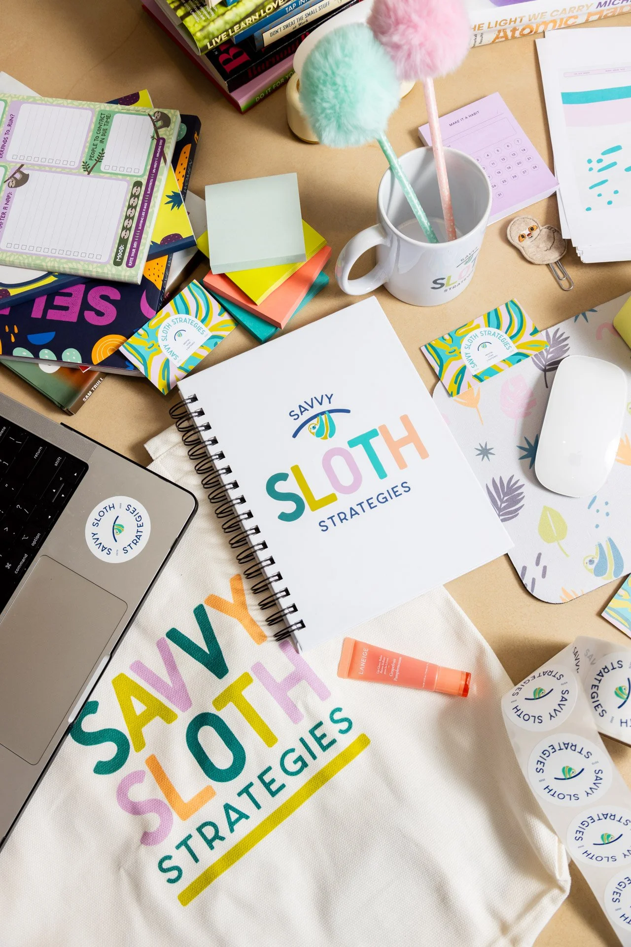 Desk with stationery and branding materials featuring 'Savvy Sloth Strategies' logo, including a notebook, mug, laptop, sticky notes, and colorful pens.