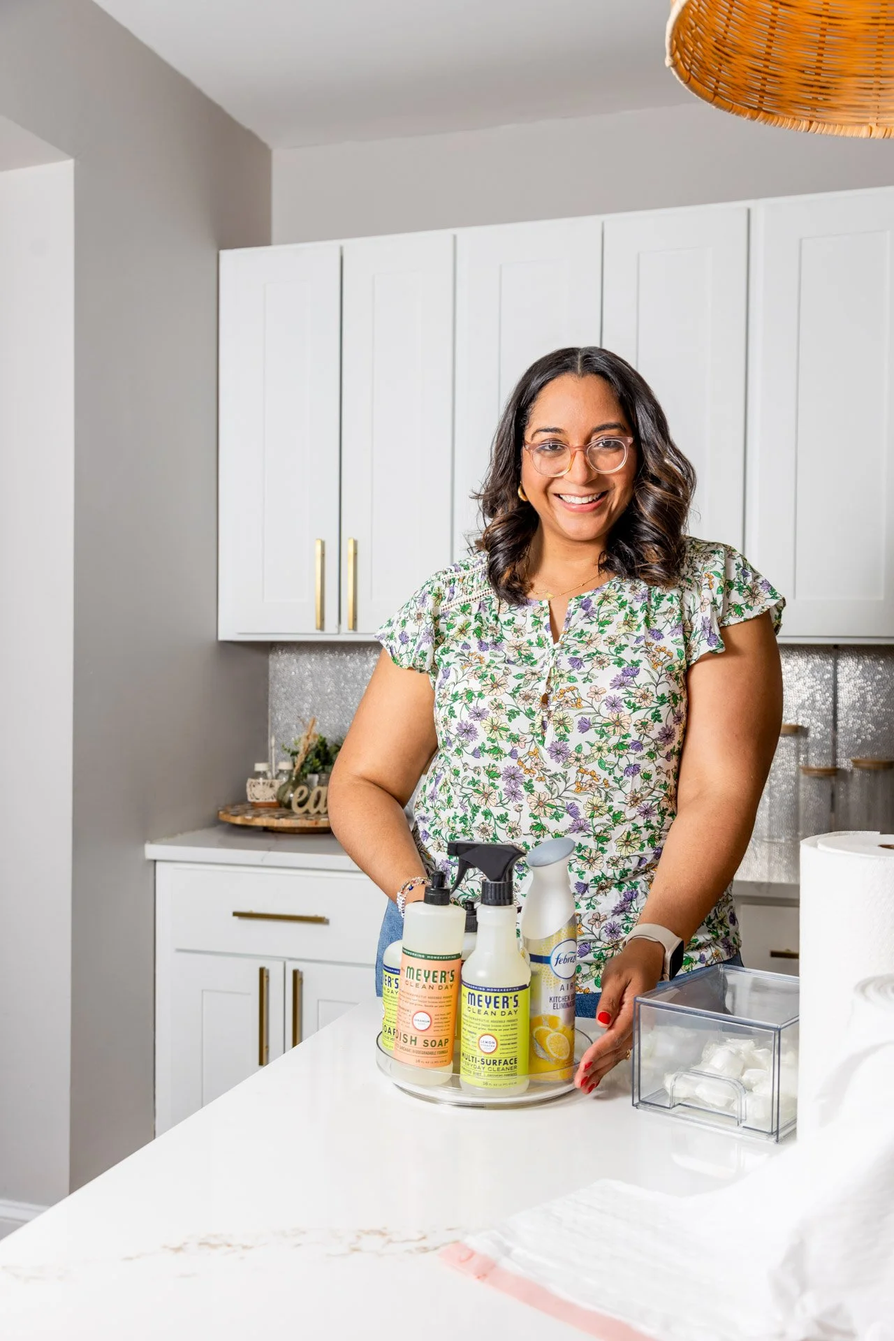 Renee smiling in a kitchen with cleaning products, including Meyer's Clean Day and Febreze, arranged on a countertop.