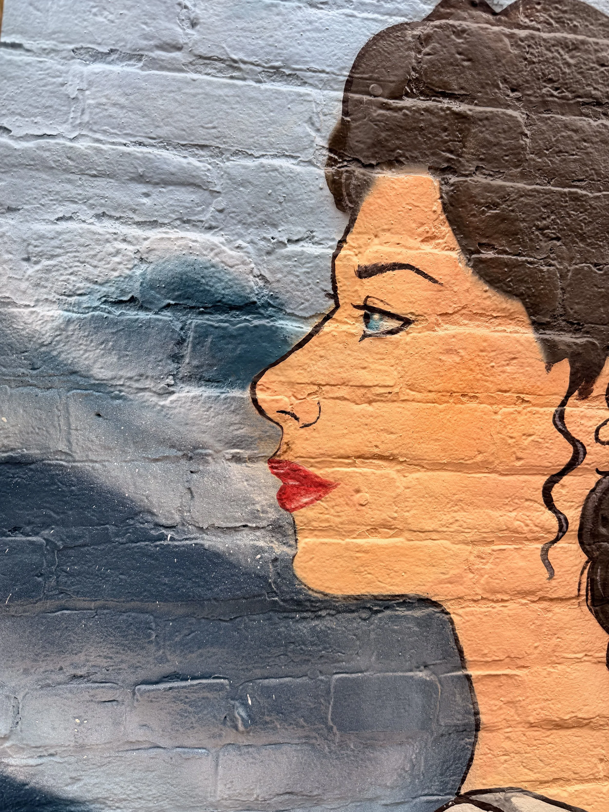 Street art mural of a woman's face with closed eyes, painted on a textured brick wall. The woman has dark hair, red lips, and a peaceful expression. The mural features both warm and cool colors.