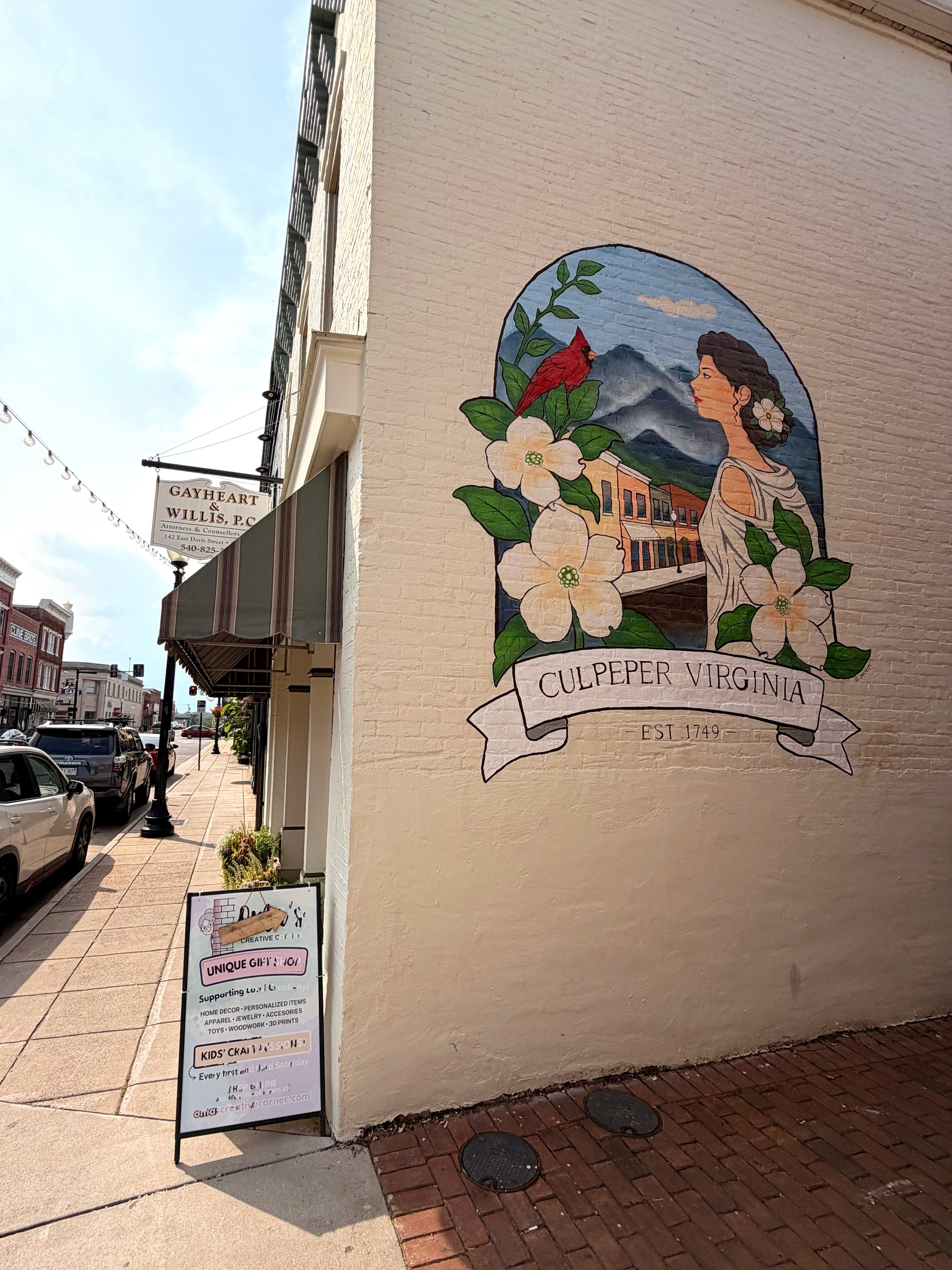 A mural on a beige brick wall depicting a woman with a flower in her hair, a red bird, flowers, mountains, and buildings, with the text "Culpeper Virginia EST. 1749." A sidewalk and street with parked cars and streetlights are visible.